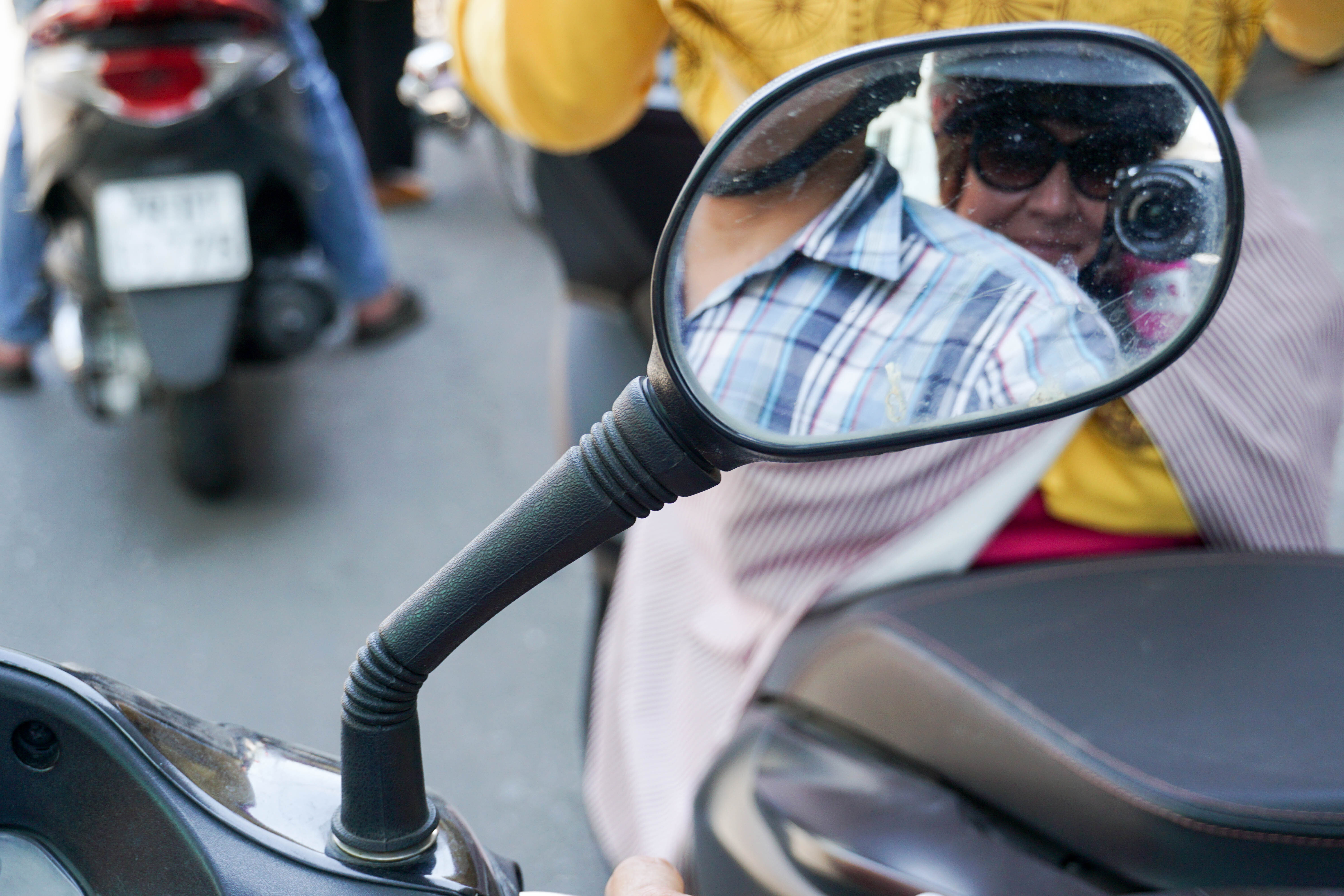 Two people, one in a yellow top and the other in a plaid shirt, are reflected in a motorcycle&#x27;s mirror, with a busy street scene in the background