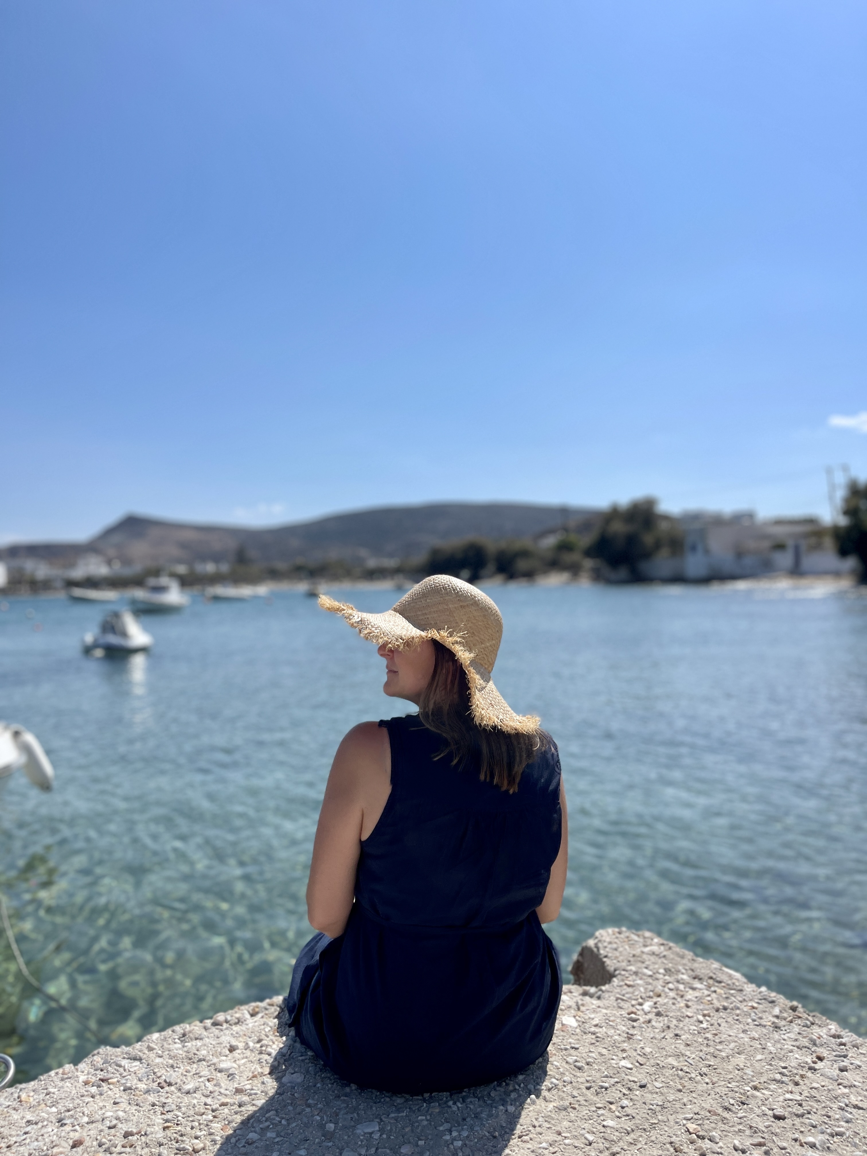 A woman in a sun hat and dark dress sits on a stone ledge by a calm body of water with boats and distant hills in the background