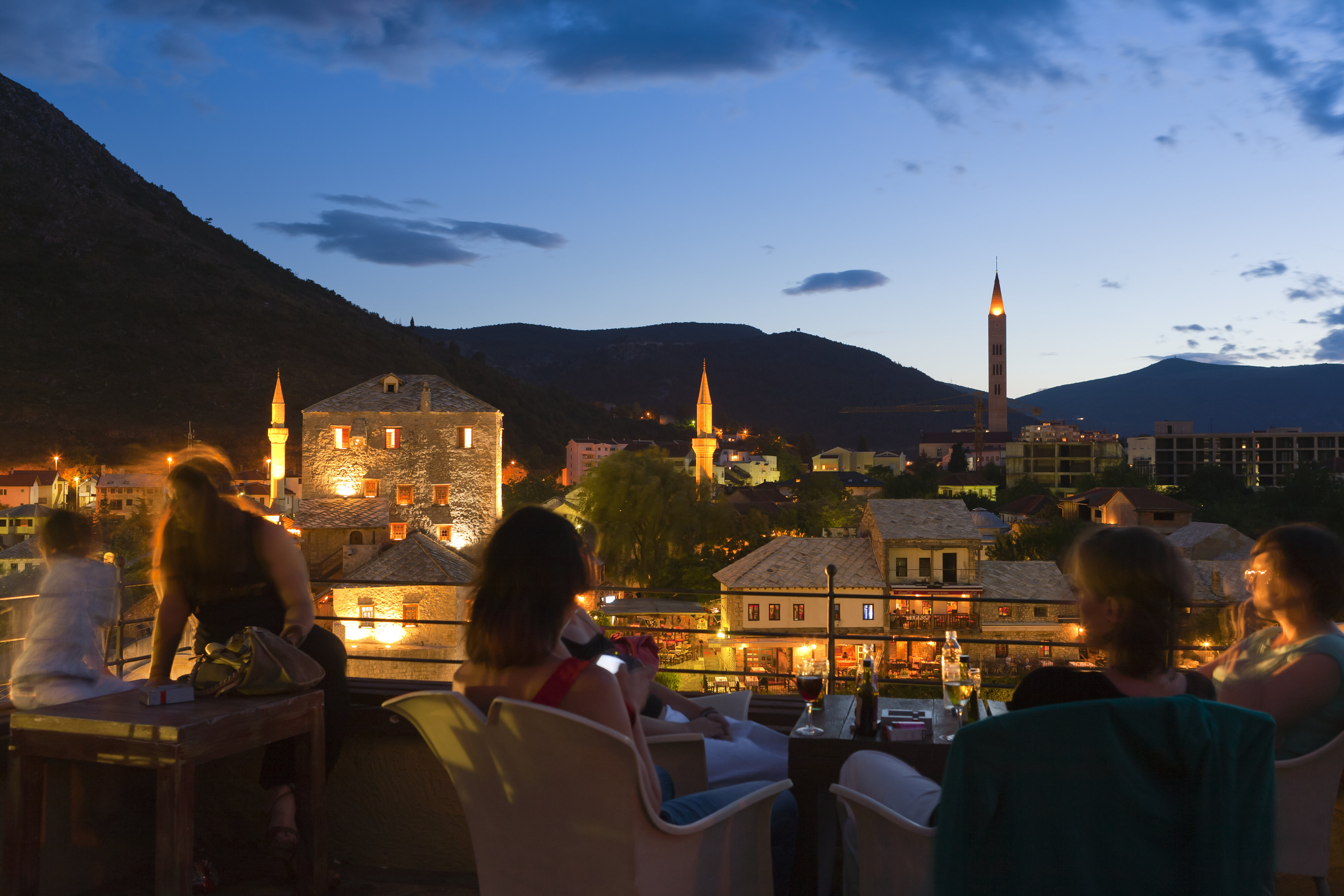 People dine on an outdoor terrace overlooking a historic town with illuminated buildings and distant mountains at dusk