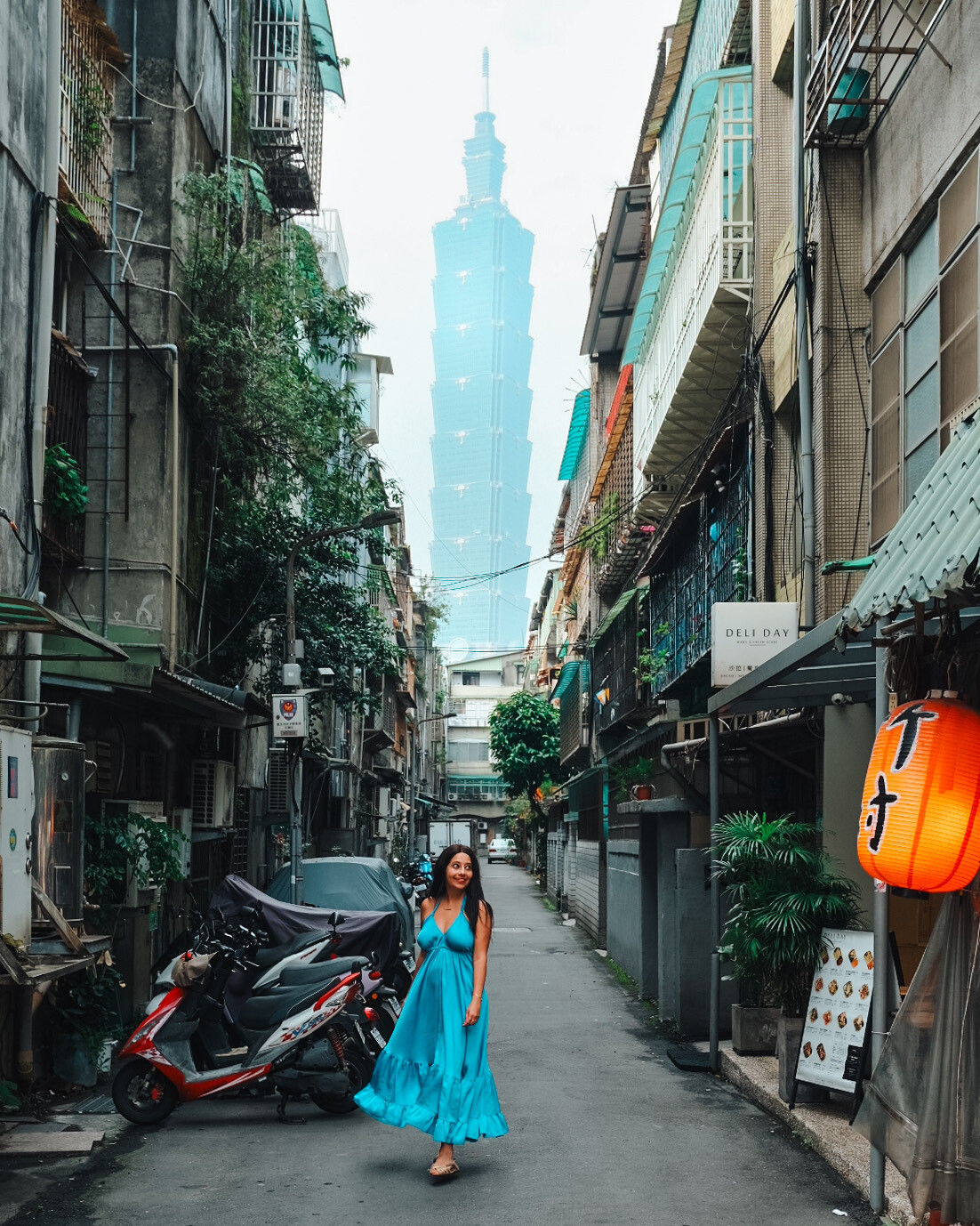 A woman in a flowing dress walks in a narrow alleyway with Taipei 101 in the background