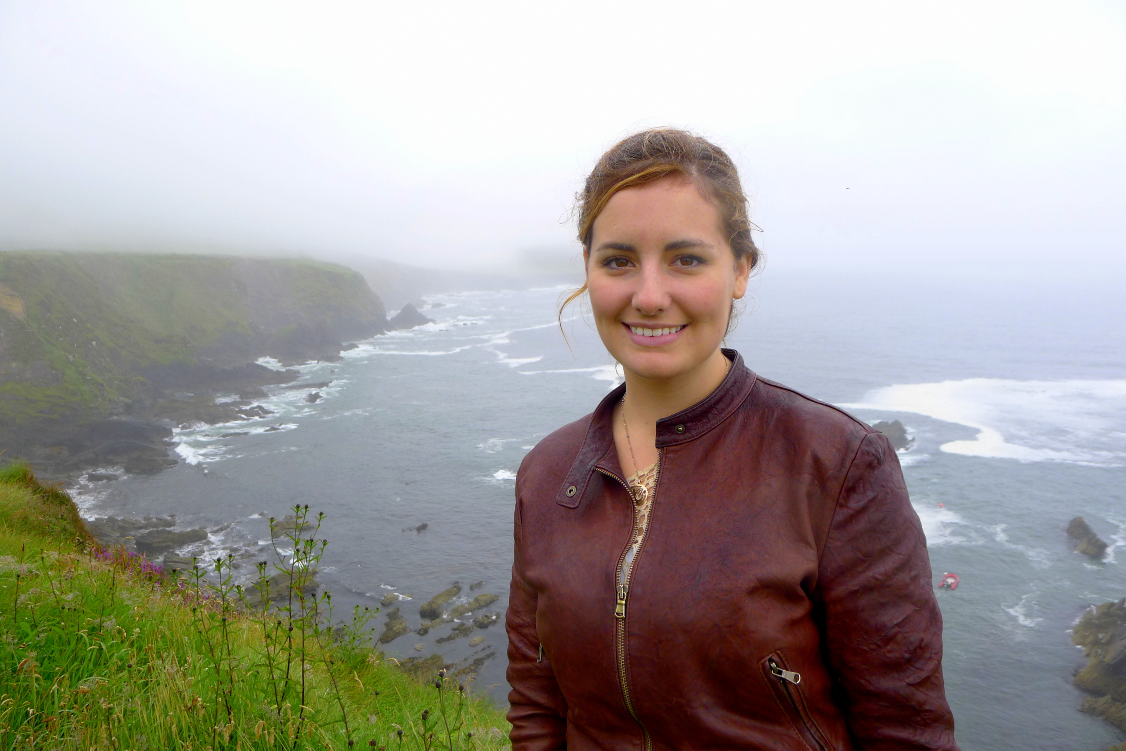 A smiling woman in a leather jacket stands in front of a foggy coastline with rocky cliffs and the ocean in the background