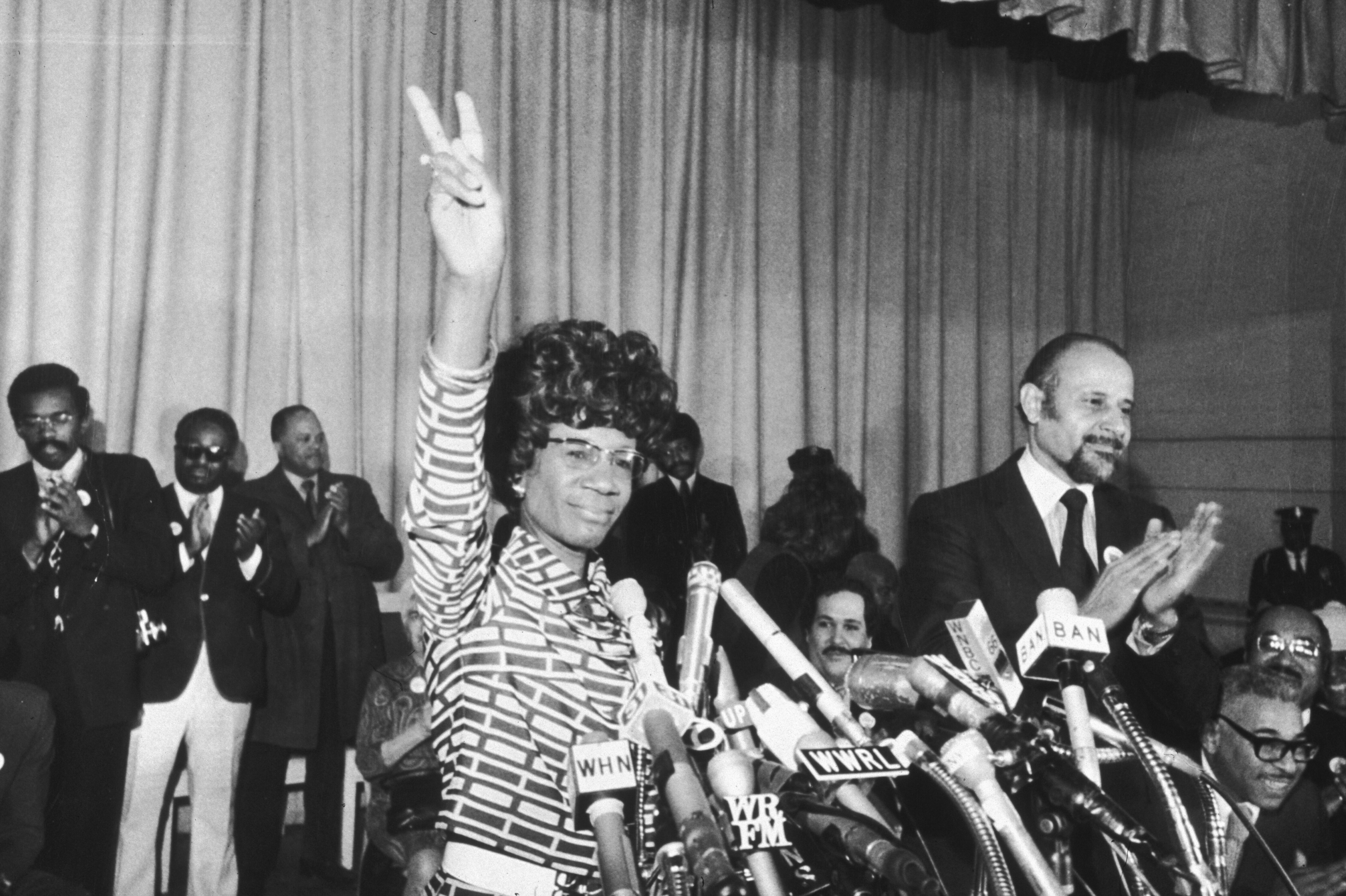 Shirley Chisholm raises a peace sign at a press conference, surrounded by microphones and supporters clapping in the background