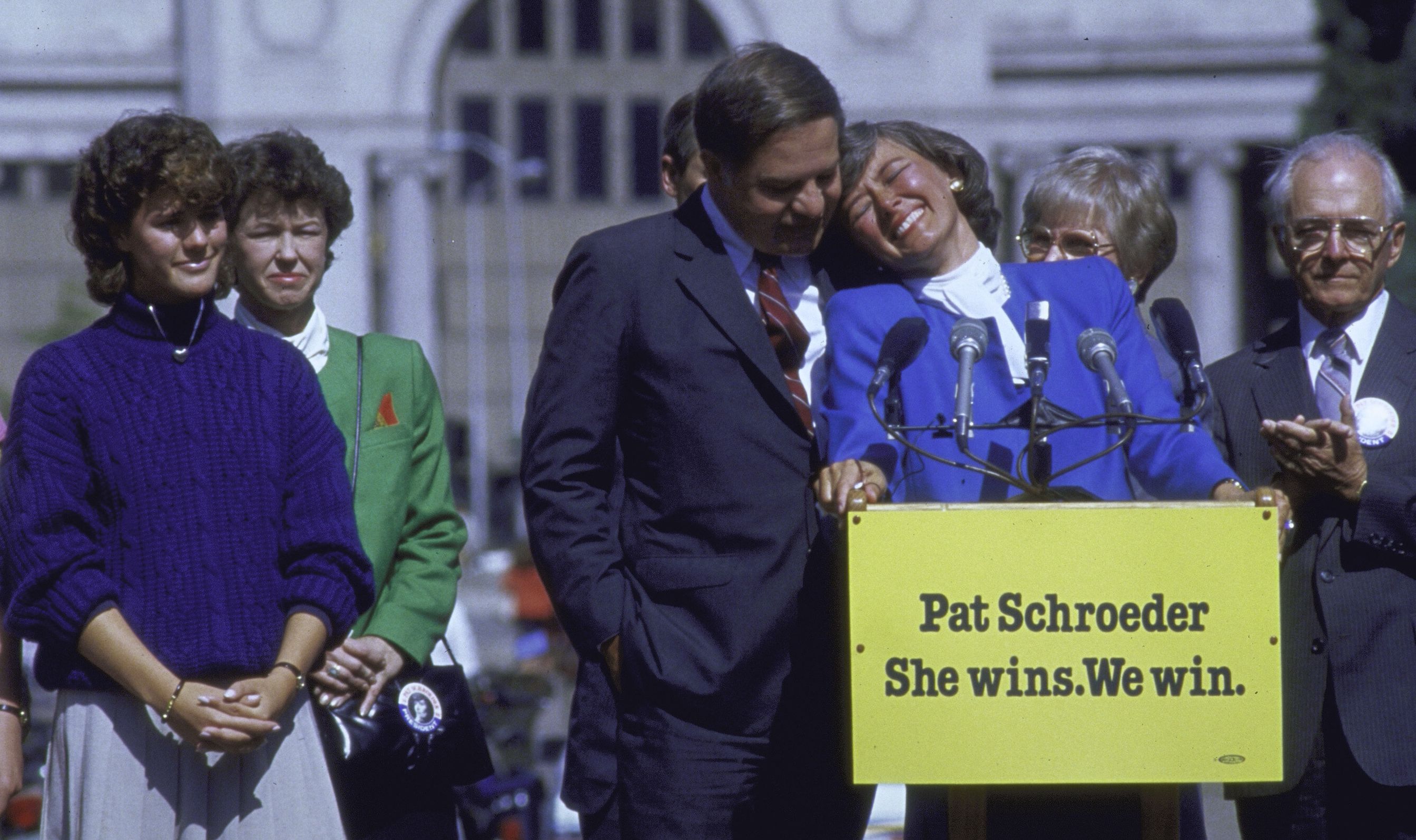 Pat Schroeder, smiling and leaning on a man's shoulder, stands at a podium with a &quot;Pat Schroeder She wins. We win.&quot; sign. Several people stand around her