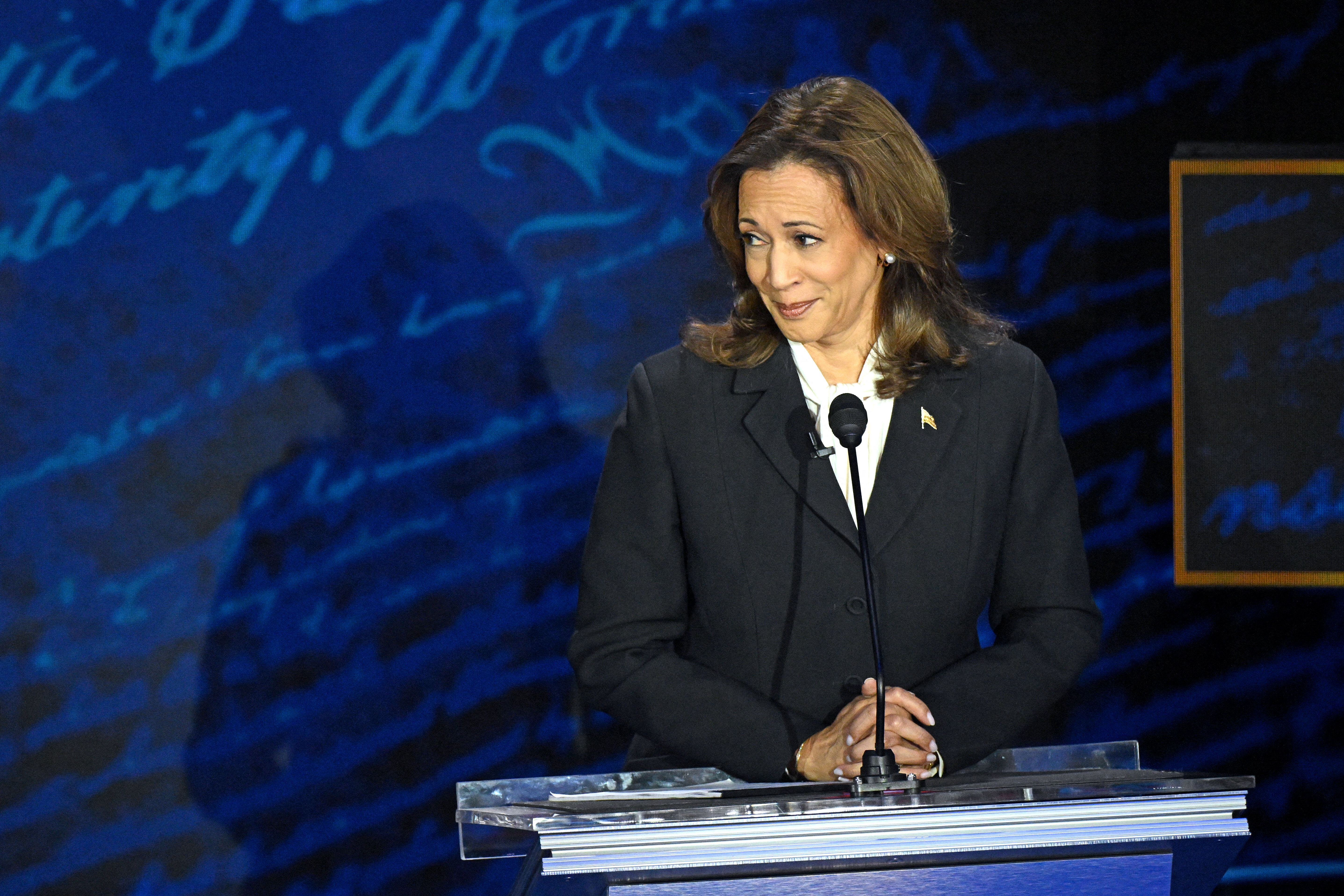 Kamala Harris stands at a podium, hands clasped, during a public speaking event. She wears a formal dark suit and a white blouse