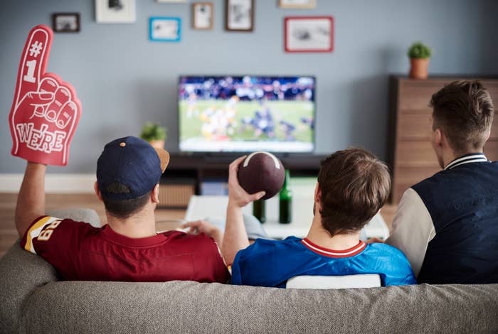 Three people sit on a couch watching a football game on TV; one holds a foam finger and another a football