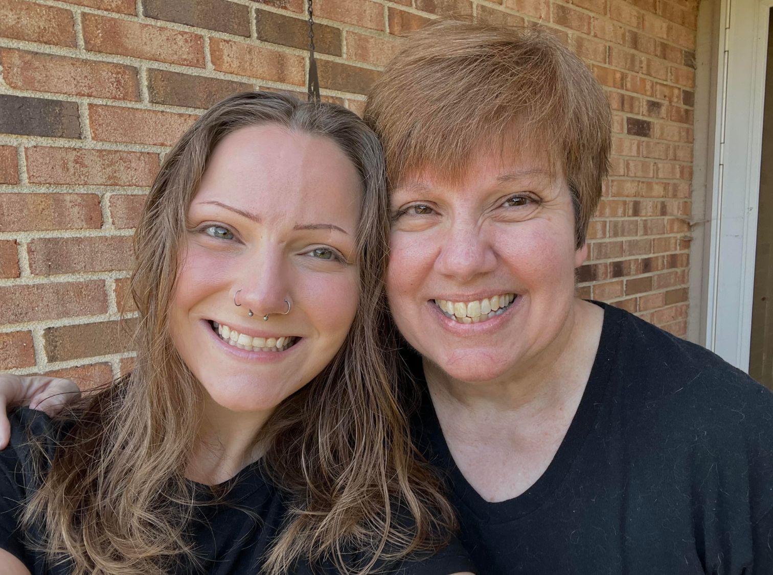 Two people smiling in front of a brick wall, both wearing black shirts
