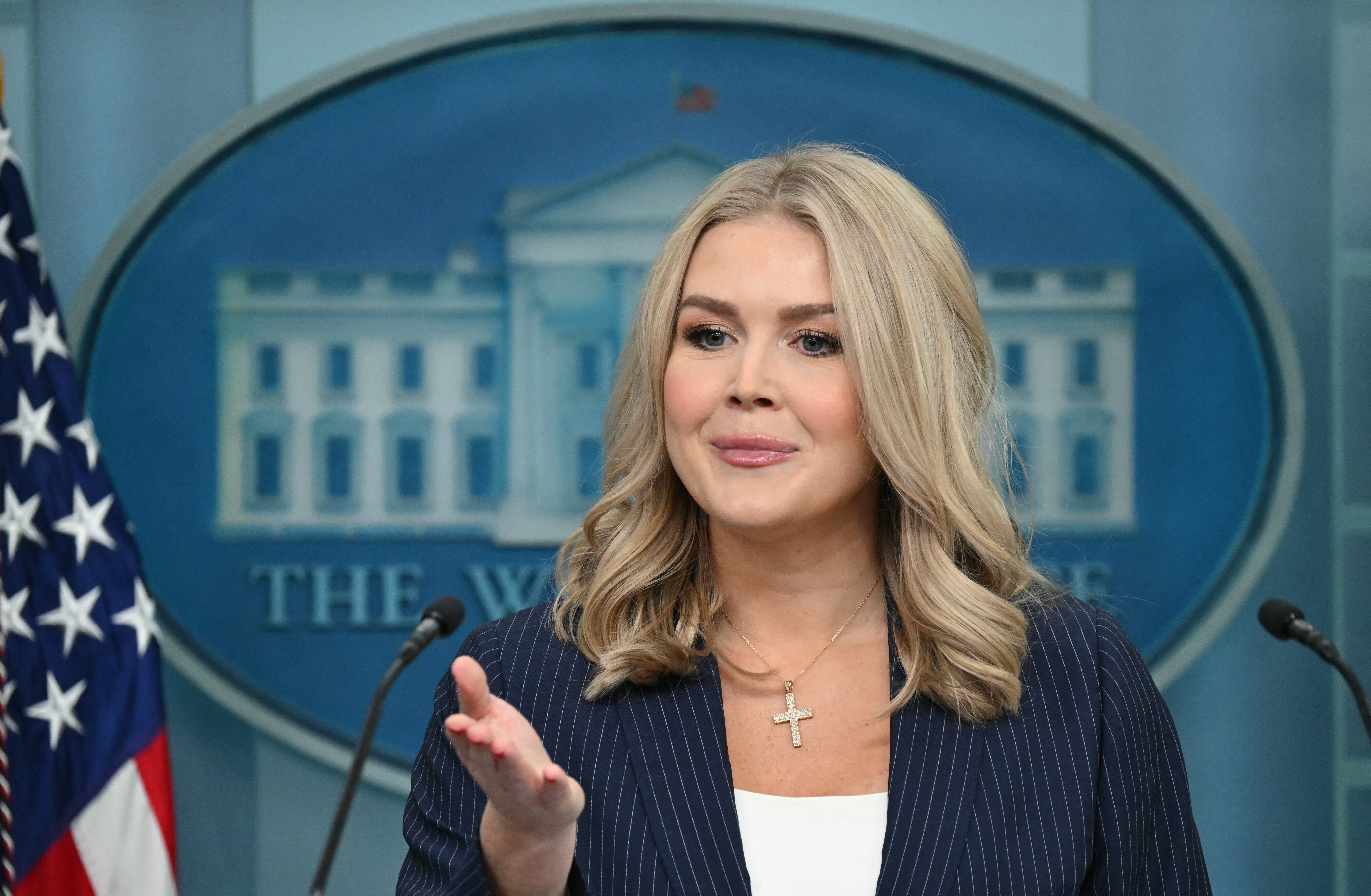 A speaker at a podium in a press briefing room, wearing a pinstripe blazer and cross necklace, gestures while addressing the audience