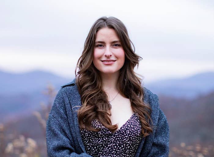 A person with long hair smiles in an outdoor setting with blurred mountains in the background. They wear a patterned outfit and a cozy cardigan