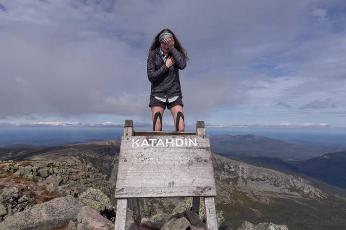 A person stands atop a mountain behind a sign reading &quot;Katahdin, Elevation 5267 Ft,&quot; enjoying the view, with a vast landscape in the background