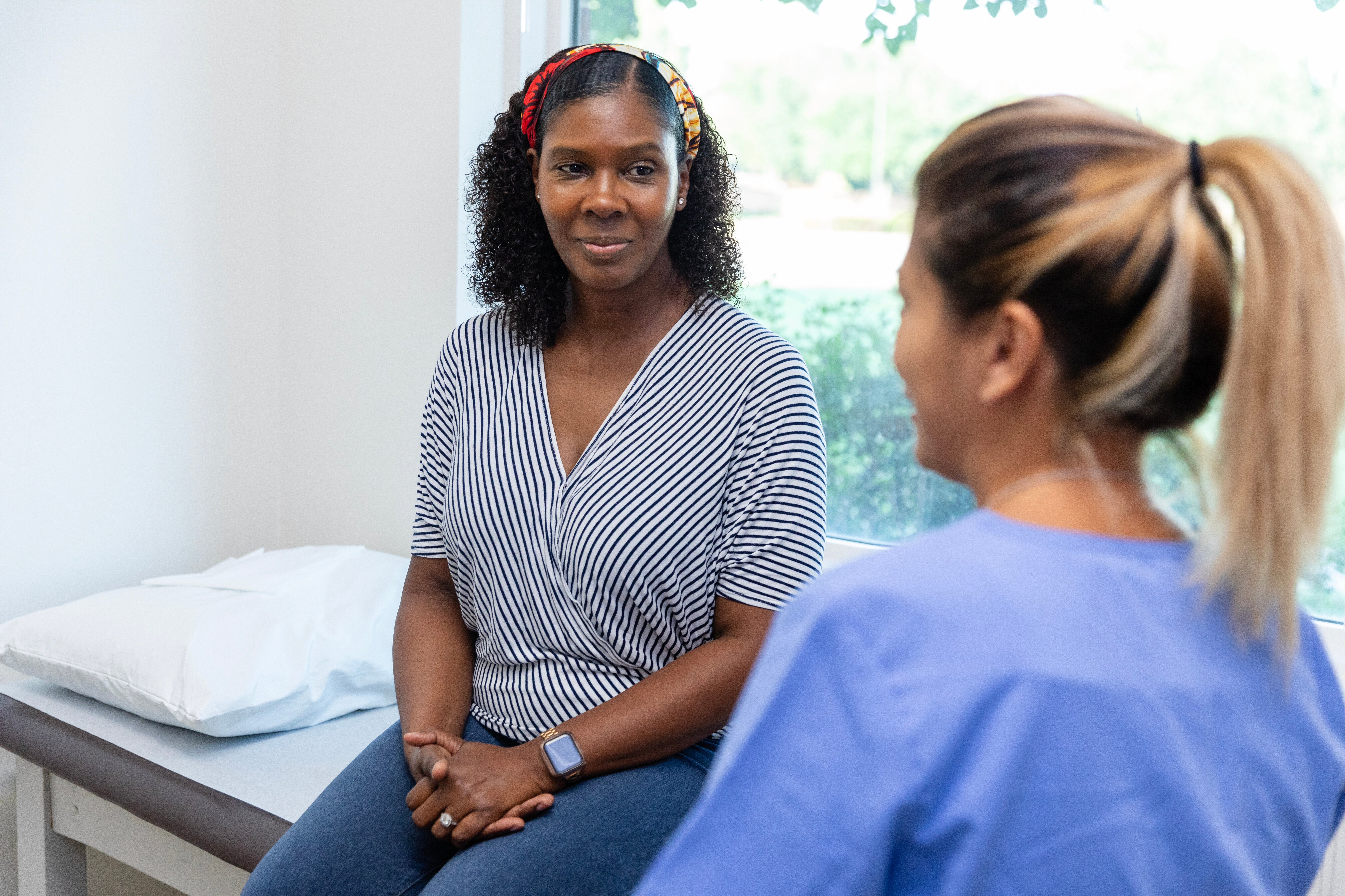 Two women sit in a medical exam room, engaging in conversation. One wears a striped top, and the other is in a medical uniform