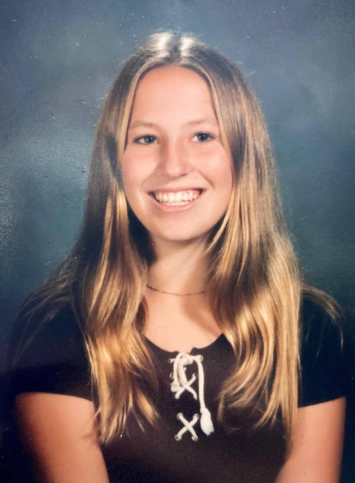 A person with long hair smiling, wearing a dark top with white criss-cross laces on the front, in a portrait setting