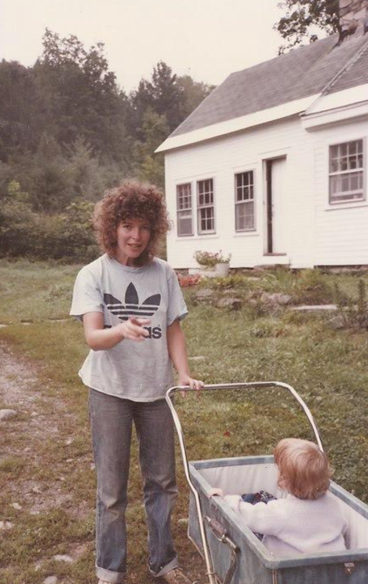 Person with curly hair, wearing a vintage Adidas shirt, stands outside a house, pointing while pushing a child in a toy wagon