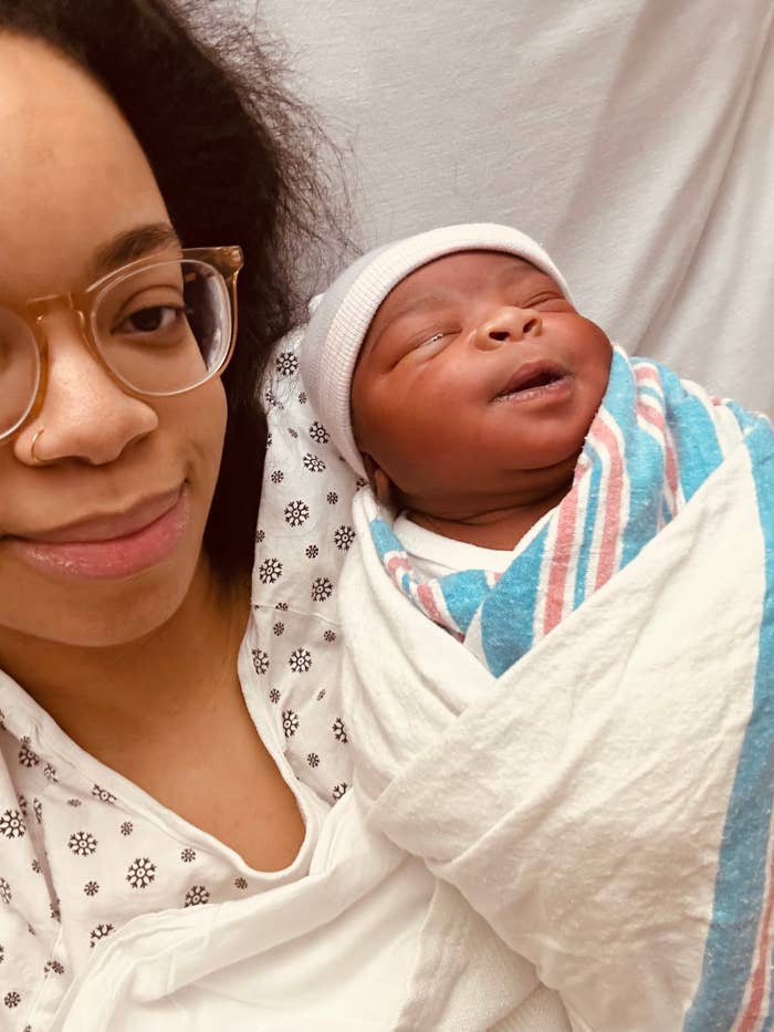 A person wearing glasses and a patterned top smiles softly while holding a newborn wrapped in a hospital blanket