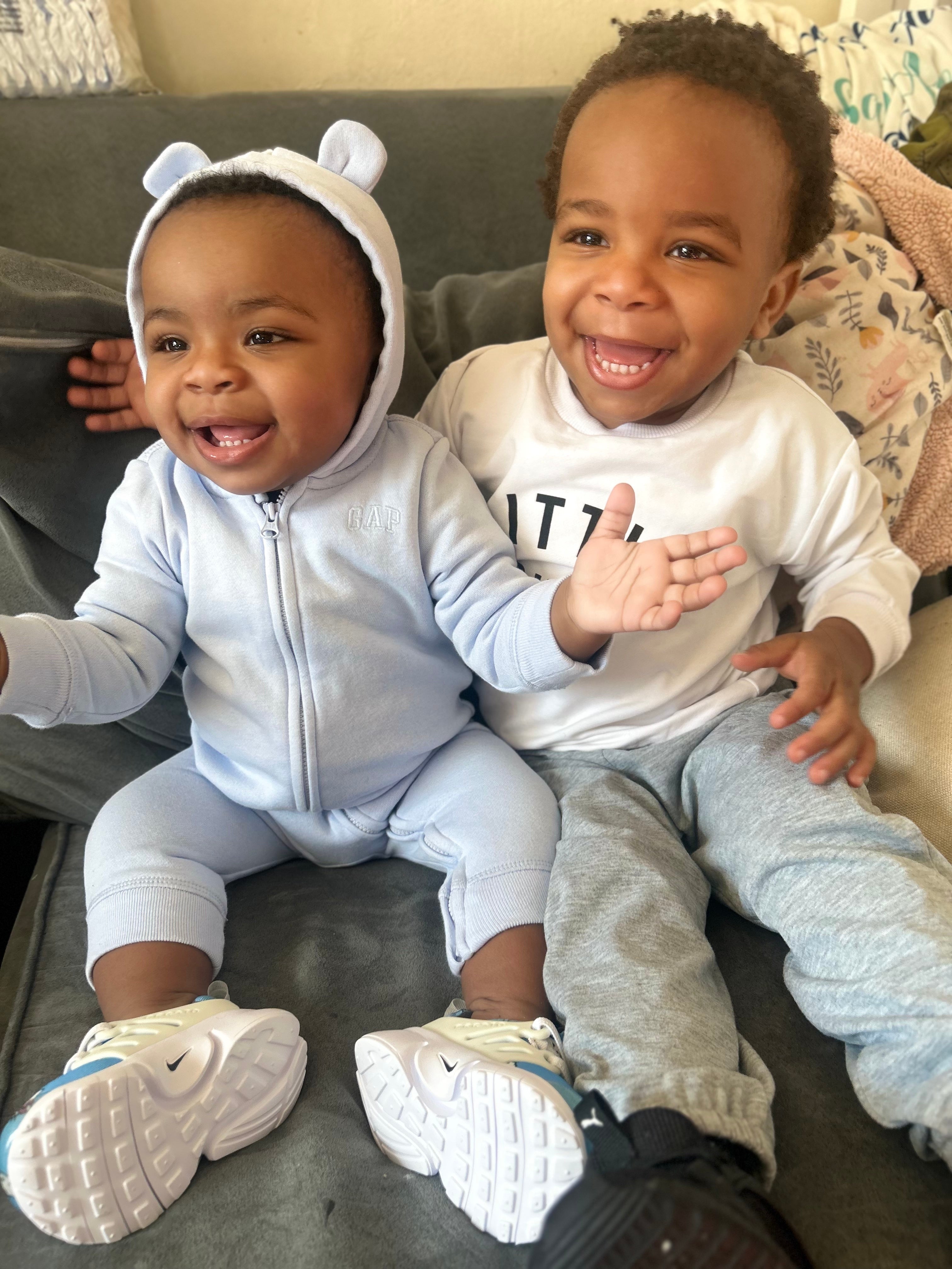 Two smiling toddlers sitting on a couch. One wears a hoodie with bear ears; the other wears a printed shirt and sweatpants