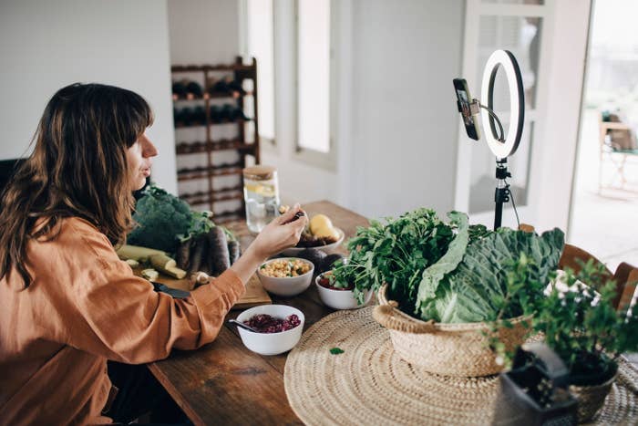 Person seated at a table filming, surrounded by various fresh vegetables, using a smartphone and ring light setup, possibly creating food content