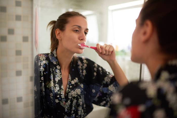A person in a patterned robe brushes their teeth in front of a bathroom mirror, reflecting a self-care routine
