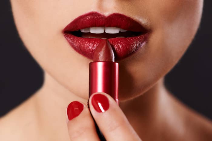 Close-up of a person applying lipstick, with focus on red lips and nail polish, holding a lipstick tube