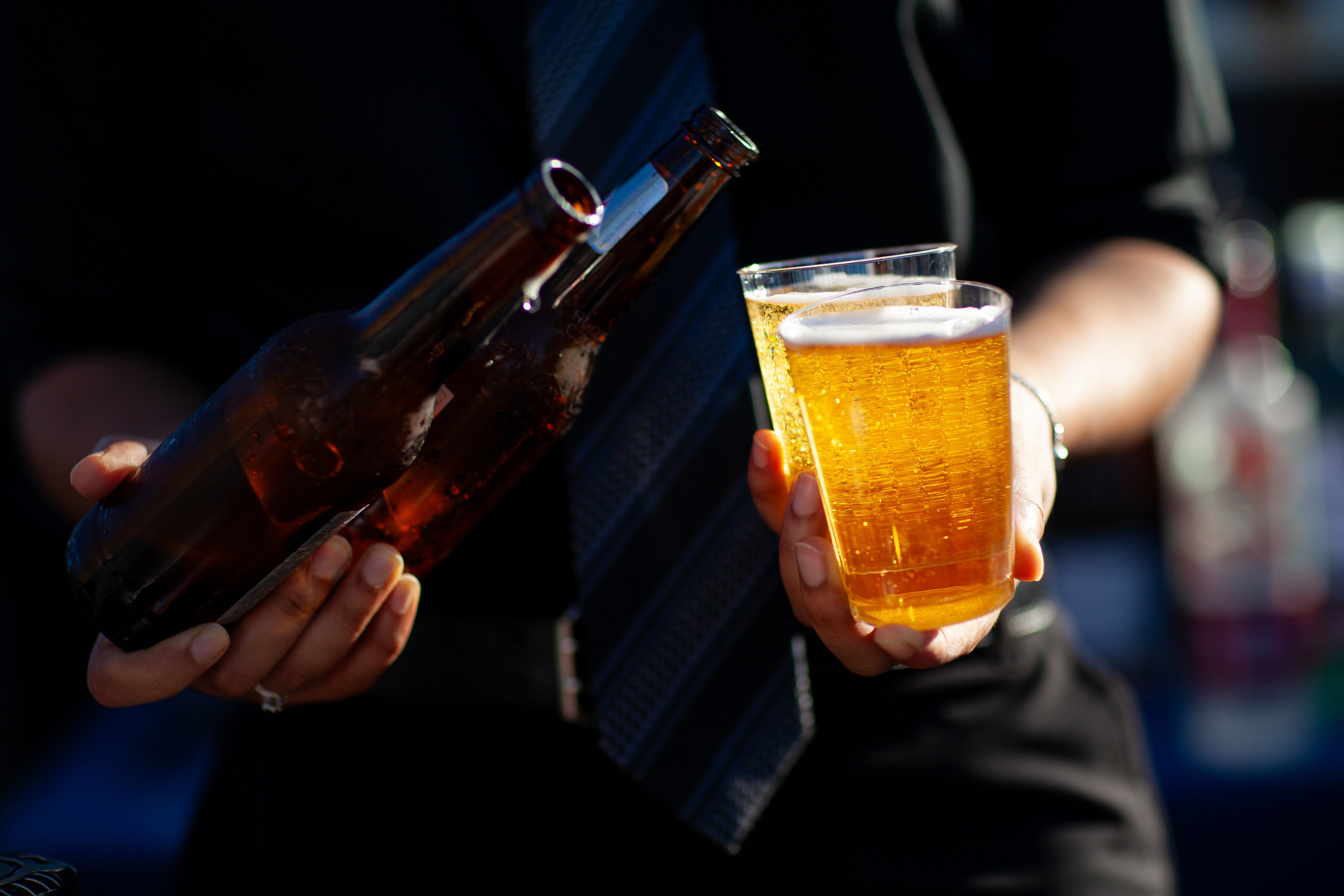 Person pouring beer from a bottle into a plastic cup, partially filled cup in the other hand, wearing a suit and tie