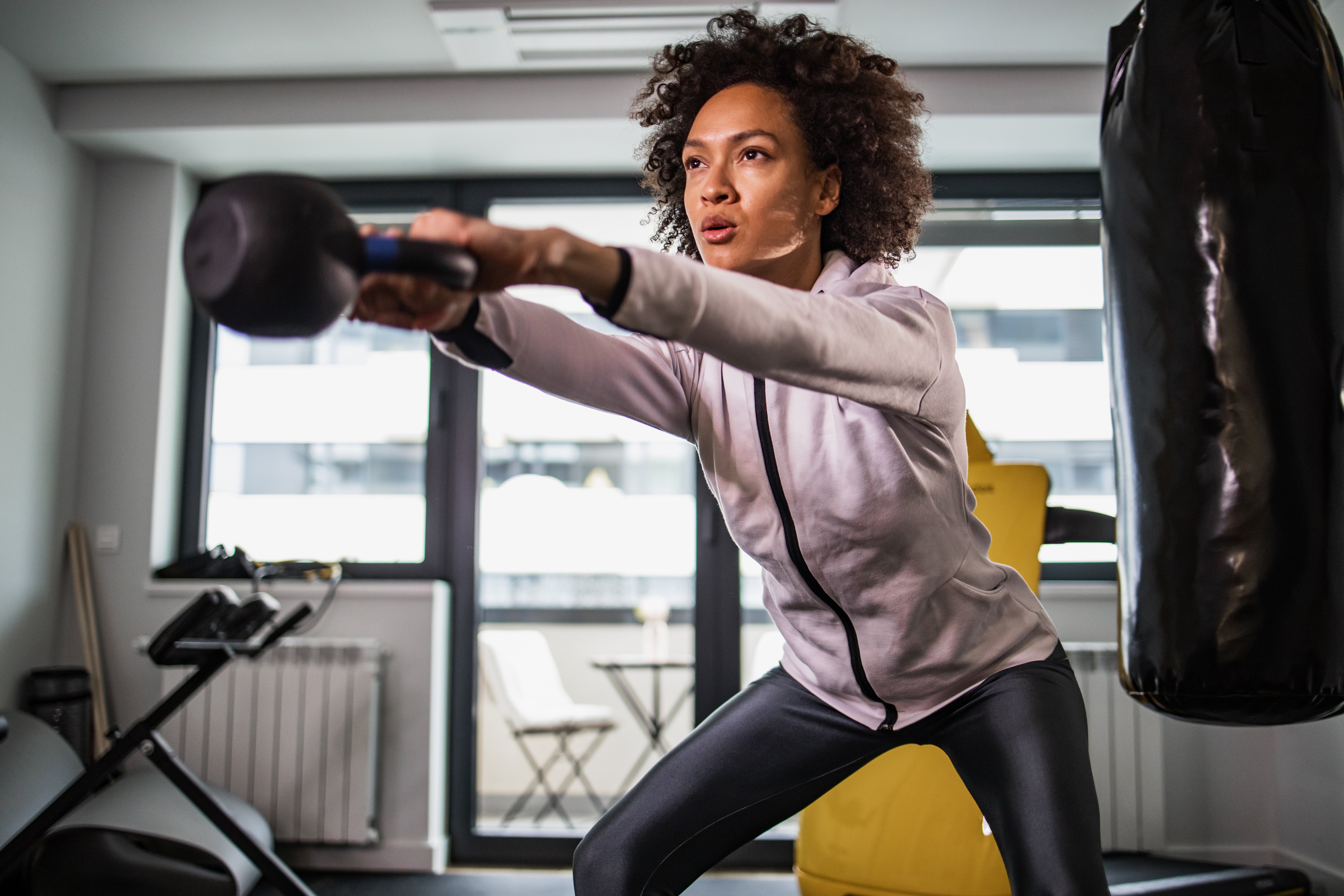 Person intensely focused, swinging kettlebell in fitness studio, wearing athletic wear with a punching bag nearby