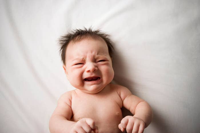 A newborn baby crying while lying on a soft surface. The baby has a tuft of dark hair and is visibly expressing discomfort or distress
