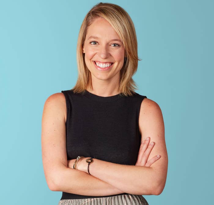 Person smiling with arms crossed, wearing a sleeveless top and patterned skirt, against a plain background