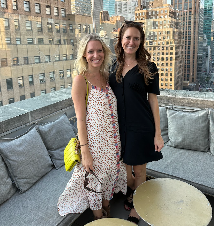 Two people smiling on a rooftop with city buildings in the background. One wears a patterned dress, the other a black dress