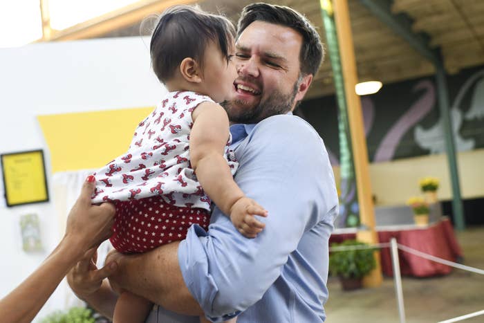 JD Vance holds a smiling baby wearing a patterned dress, creating a warm, playful moment in an indoor setting