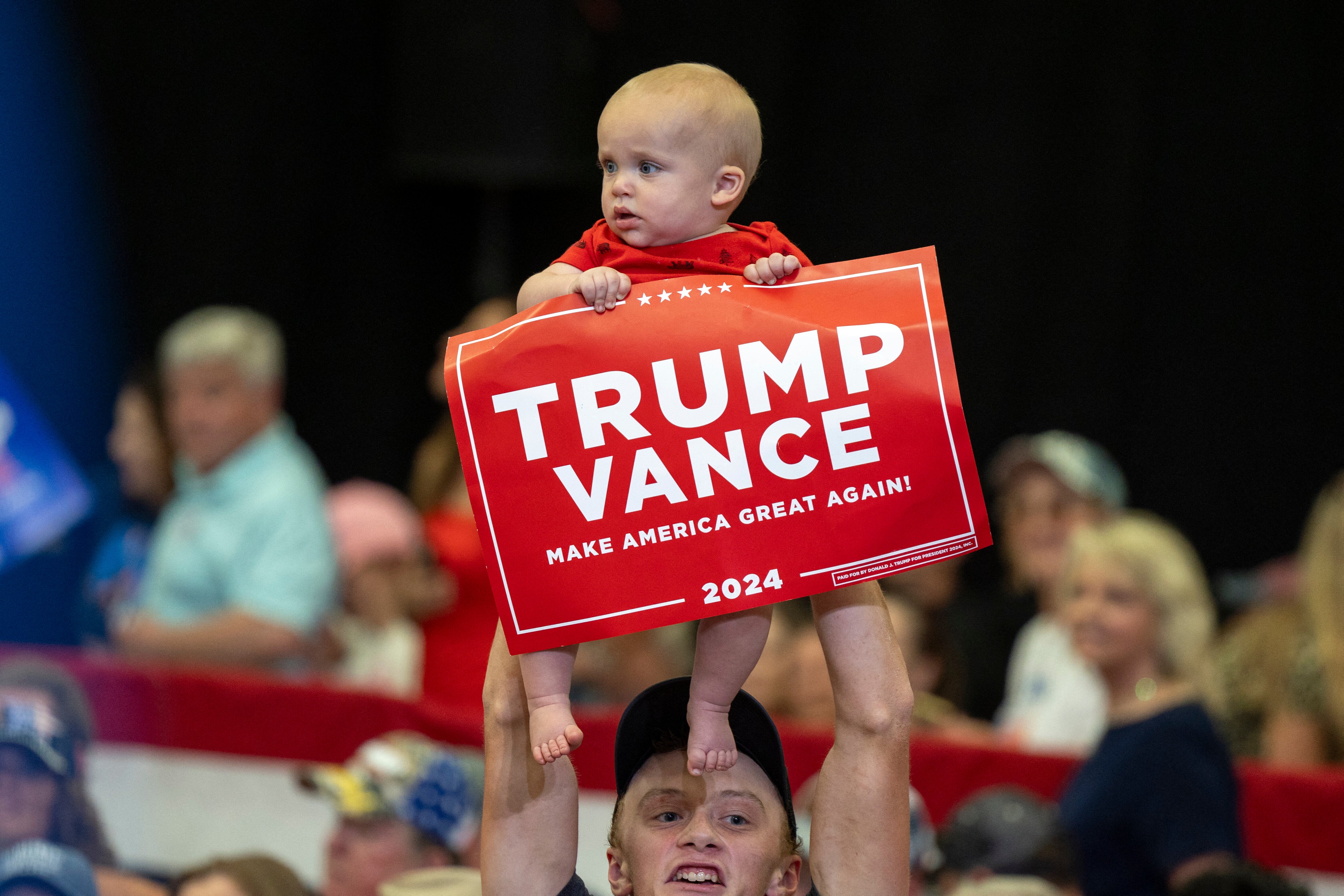 A baby held up by an adult holds a &quot;Trump Vance 2024&quot; sign at a public event