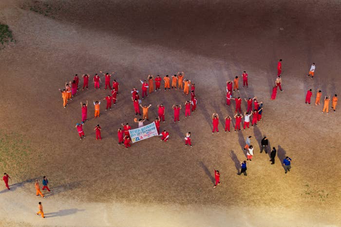 Inmates in red and orange form the word &quot;SOS&quot; on dirt ground, holding a sign in the center. People in dark clothing stand nearby