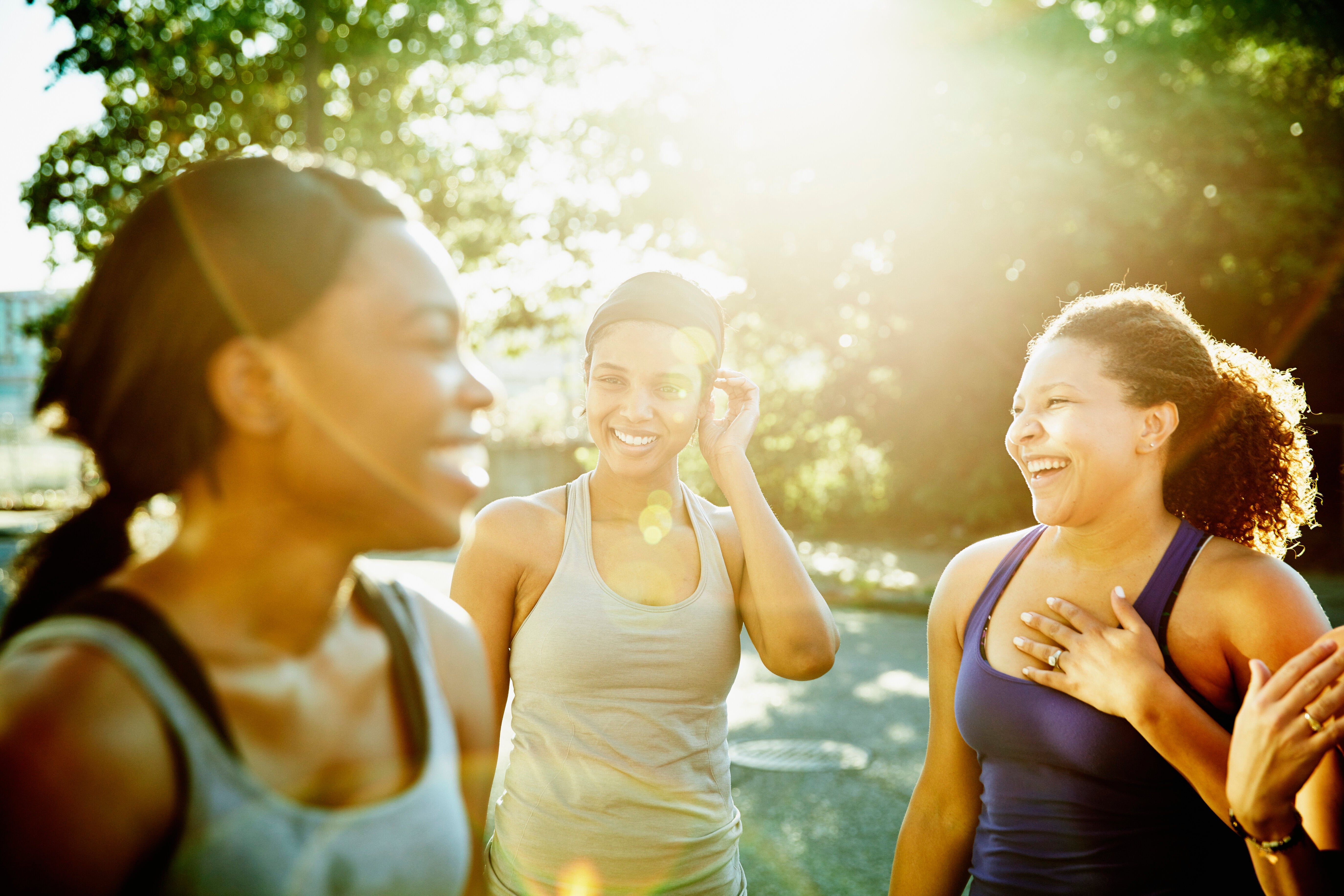 Three women in athletic wear smiling and talking outdoors in sunlight