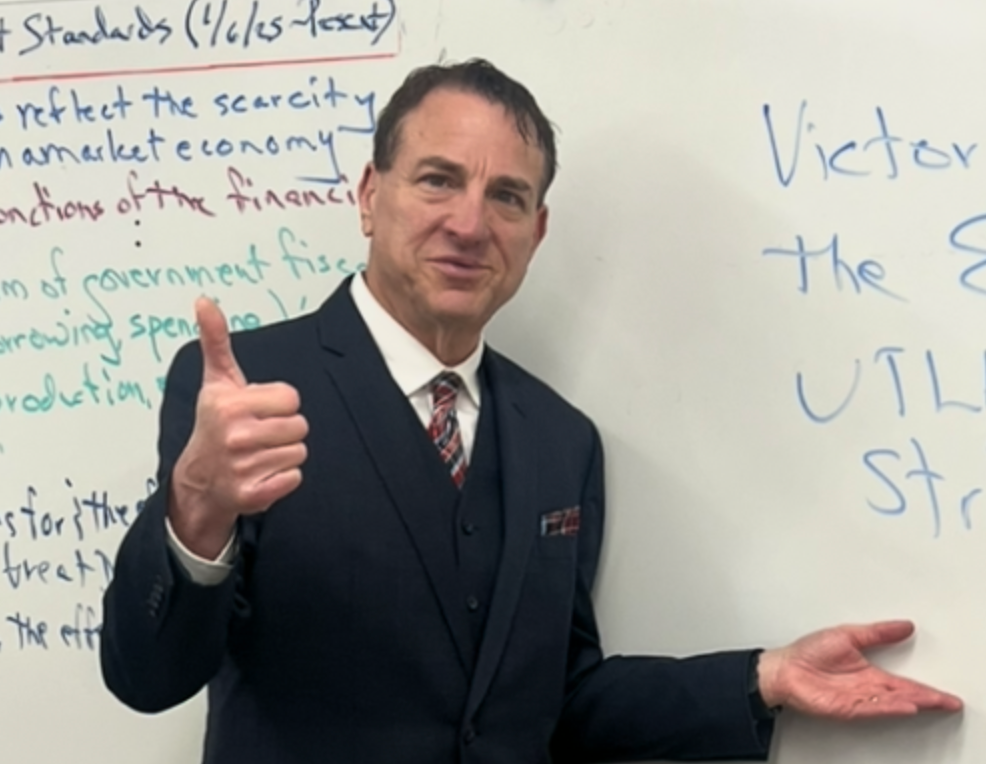 Man in a suit and tie gives a thumbs-up in front of a whiteboard with partially visible text