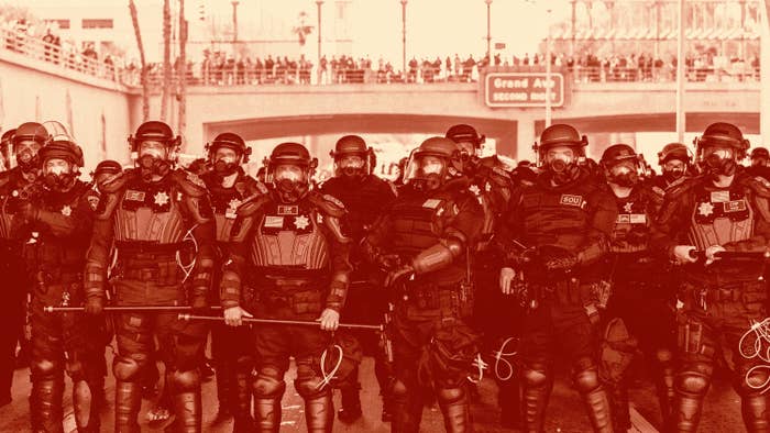 A group of uniformed officers in riot gear stand in front of a bridge, holding shields and batons, positioned in a line facing the camera