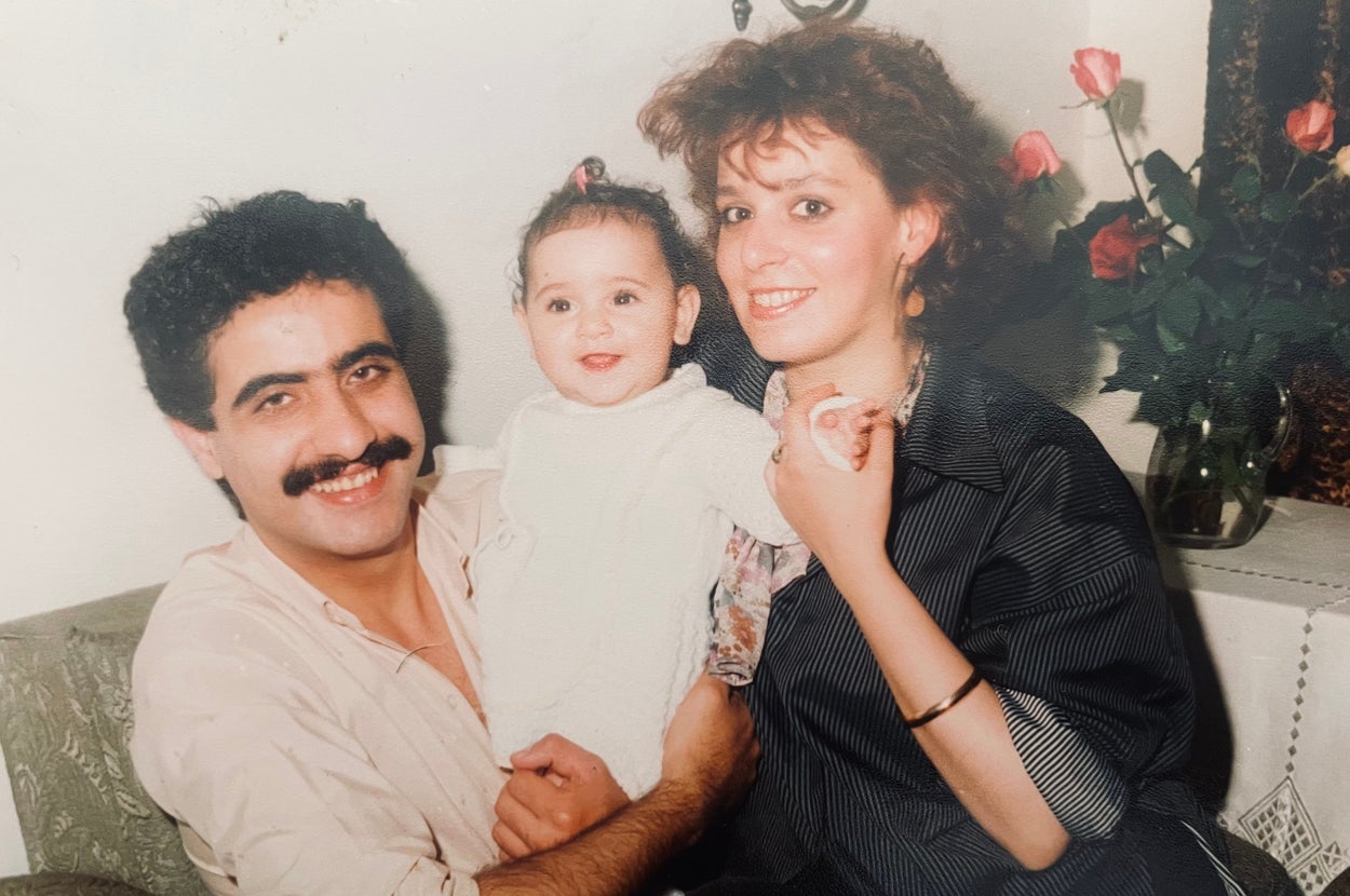 A smiling couple holds a baby between them, sitting on a couch with roses in the background