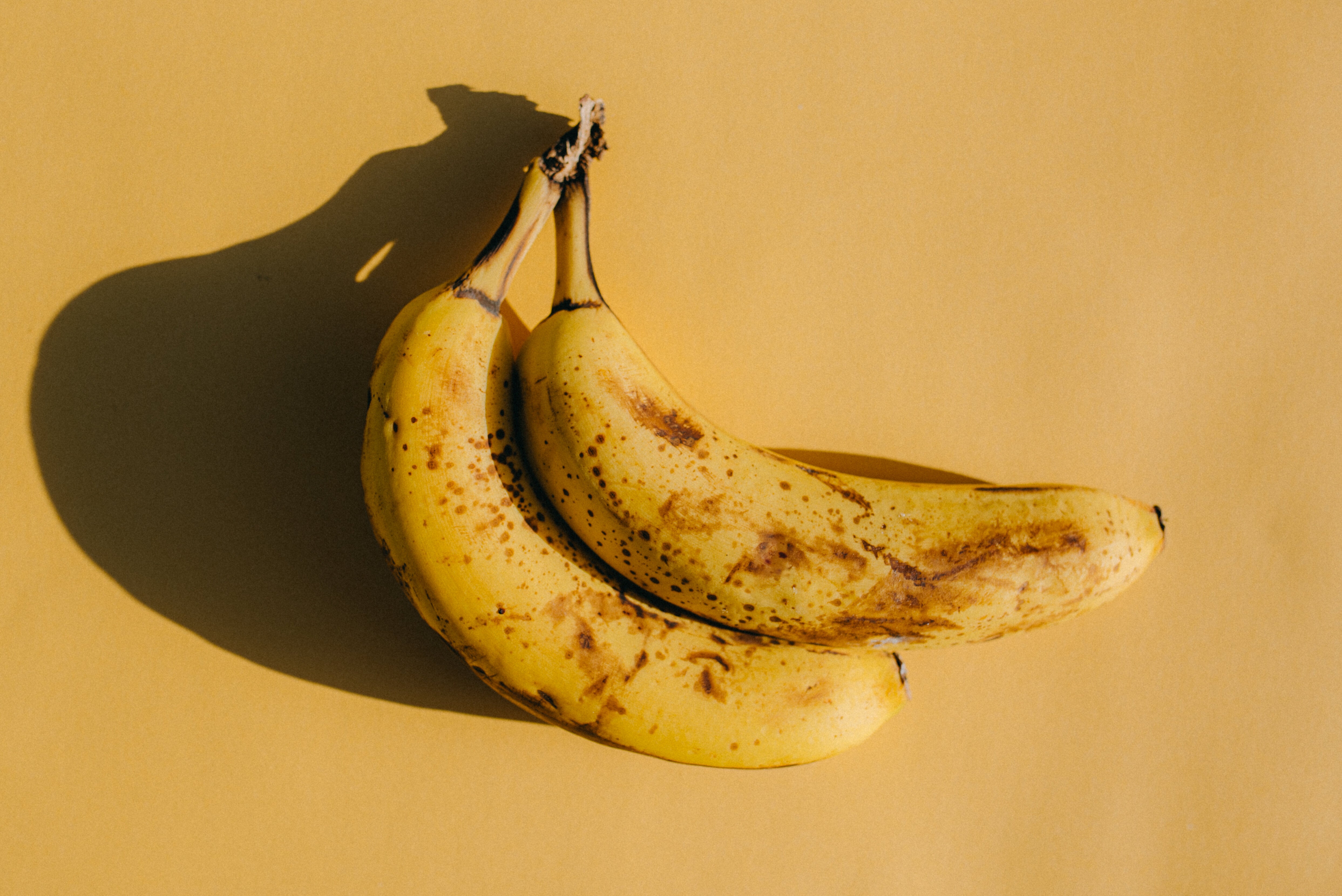 Two ripe bananas with brown spots resting together on a flat surface, casting a shadow to the left