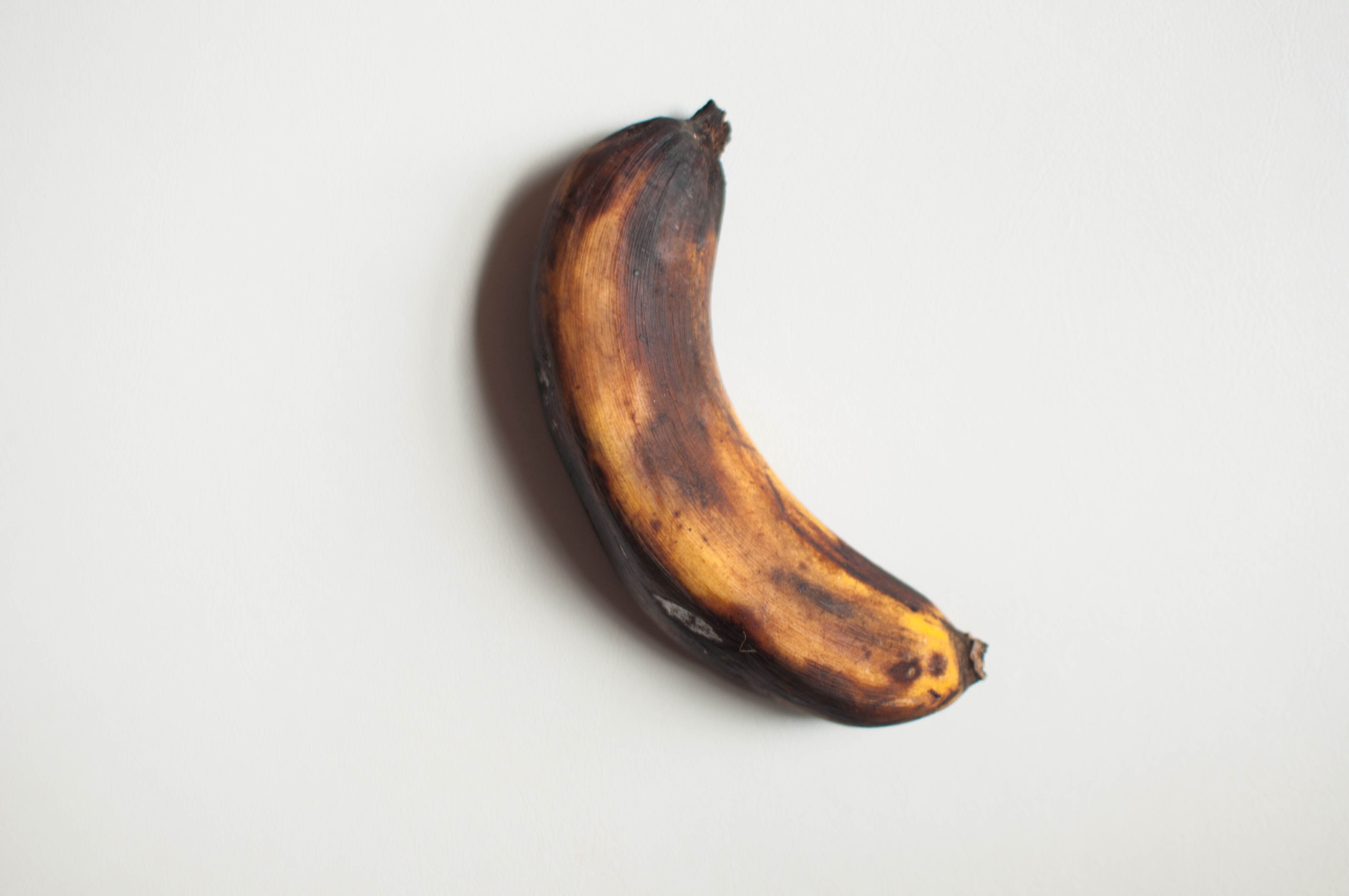 A ripe banana with a mostly brown peel, showing signs of overripeness against a plain background