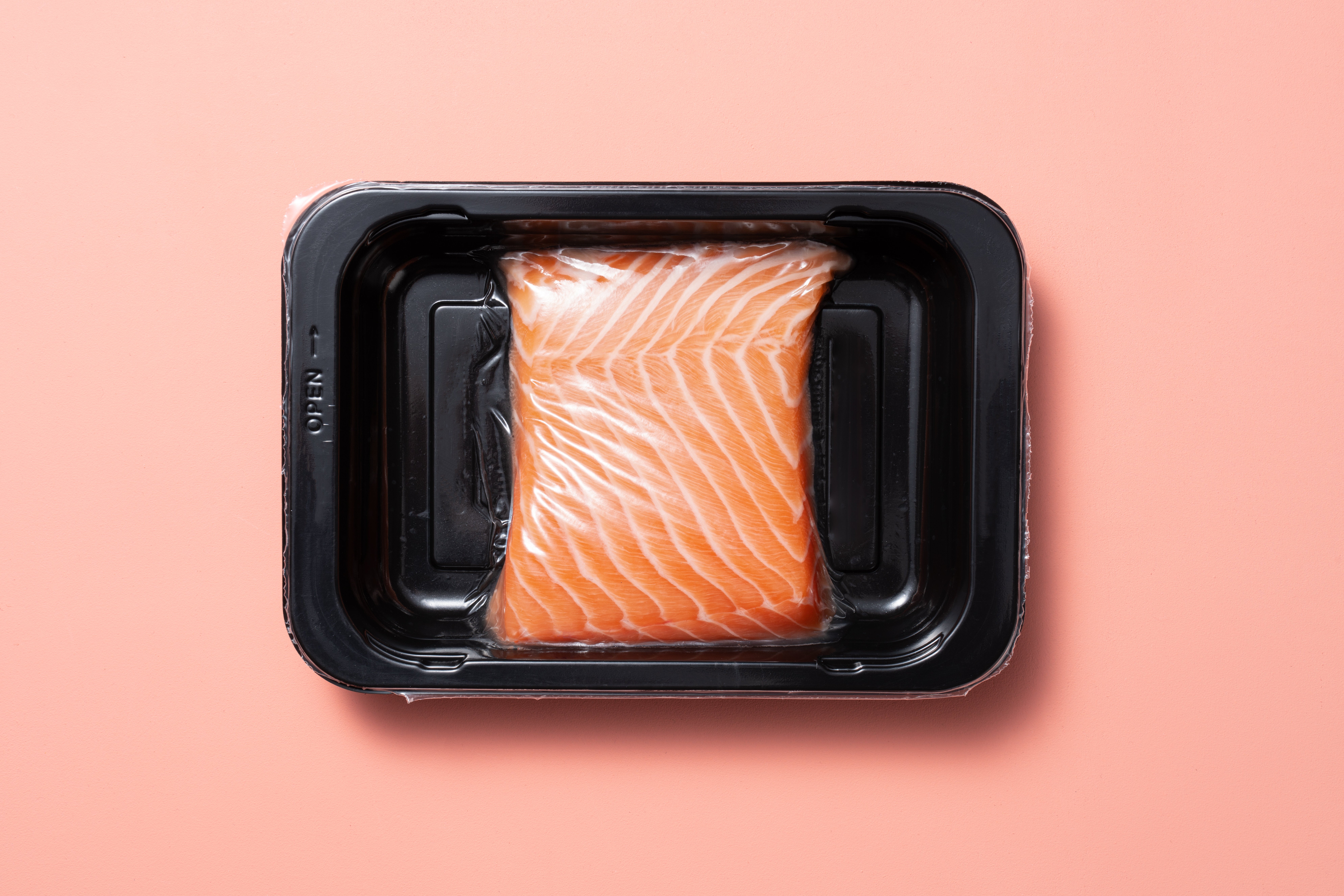 Packaged raw salmon filet in a black tray, sealed with plastic wrap, placed on a simple backdrop