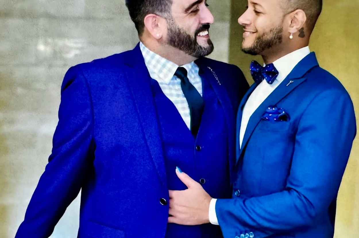 Two people in blue suits smiling at each other affectionately, with one wearing a checkered shirt and the other a bow tie