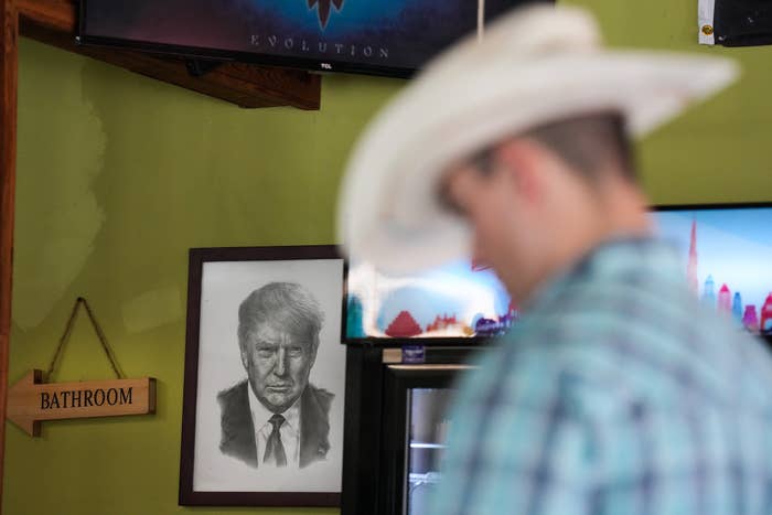 A person in a cowboy hat stands near a framed sketch of a politician and a bathroom sign, in a room with various decorations