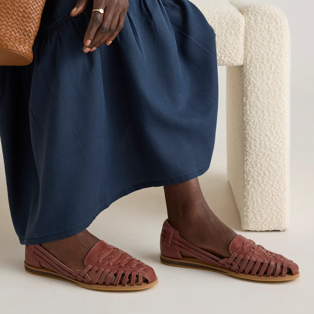 Person seated connected  a textured chair, wearing a flowing skirt and woven loafers, holding a textured bag