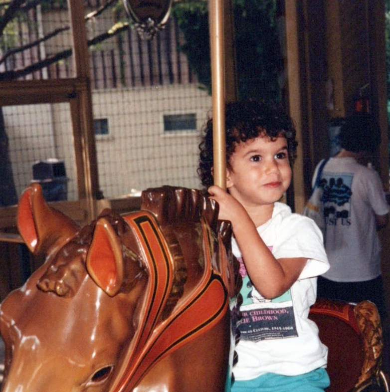Child riding a carousel horse, holding the pole and smiling, in an indoor setting