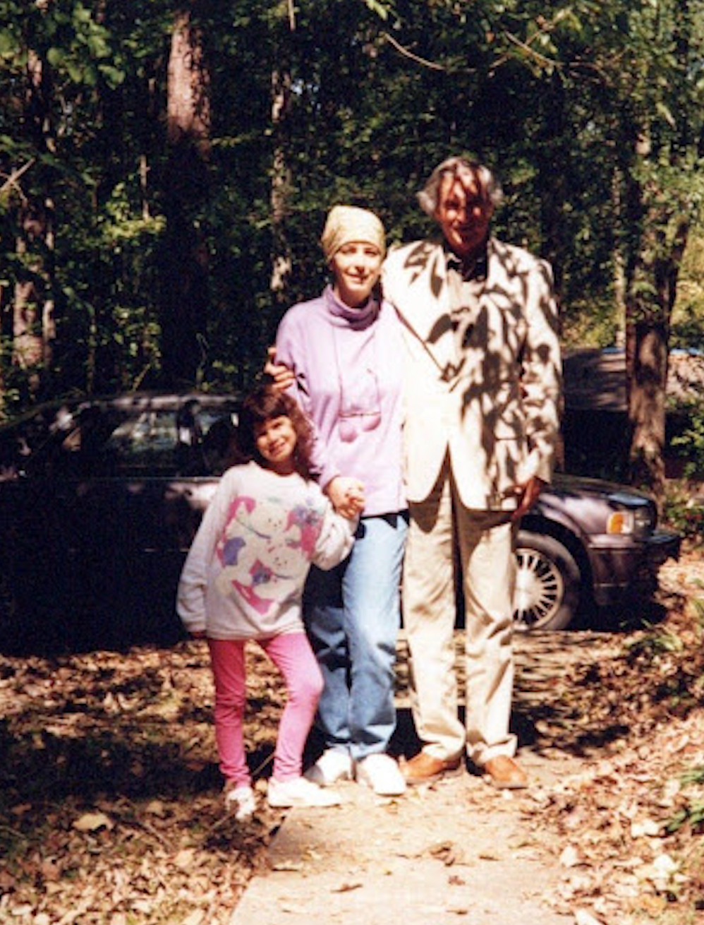 Three people stand outdoors on a leafy path. An older man in a light suit, a woman in a casual outfit, and a child pose together