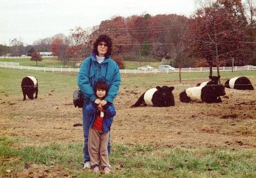 A person and child smile in front of grazing cows in a pasture with trees and a fence in the background