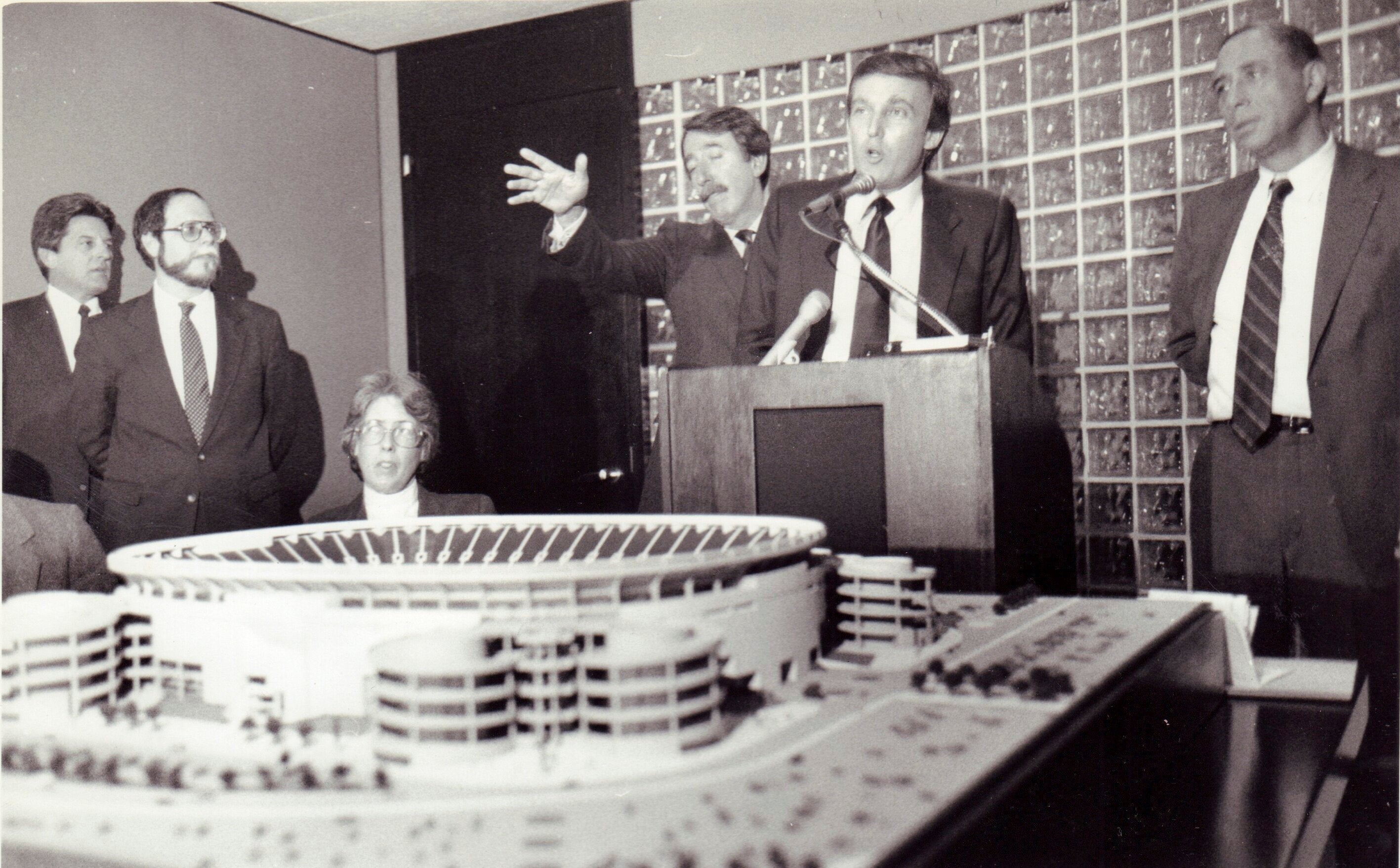 Group of 5 men successful suits and 1 woman, gathered astir a stadium model. One antheral astatine a podium speaks, gesturing widely