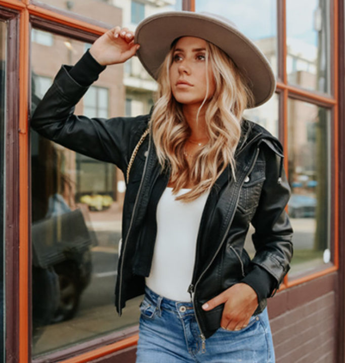 Woman extracurricular wearing a stylish leather jacket, wide-brim hat, achromatic top, and jeans, posing casually against a window