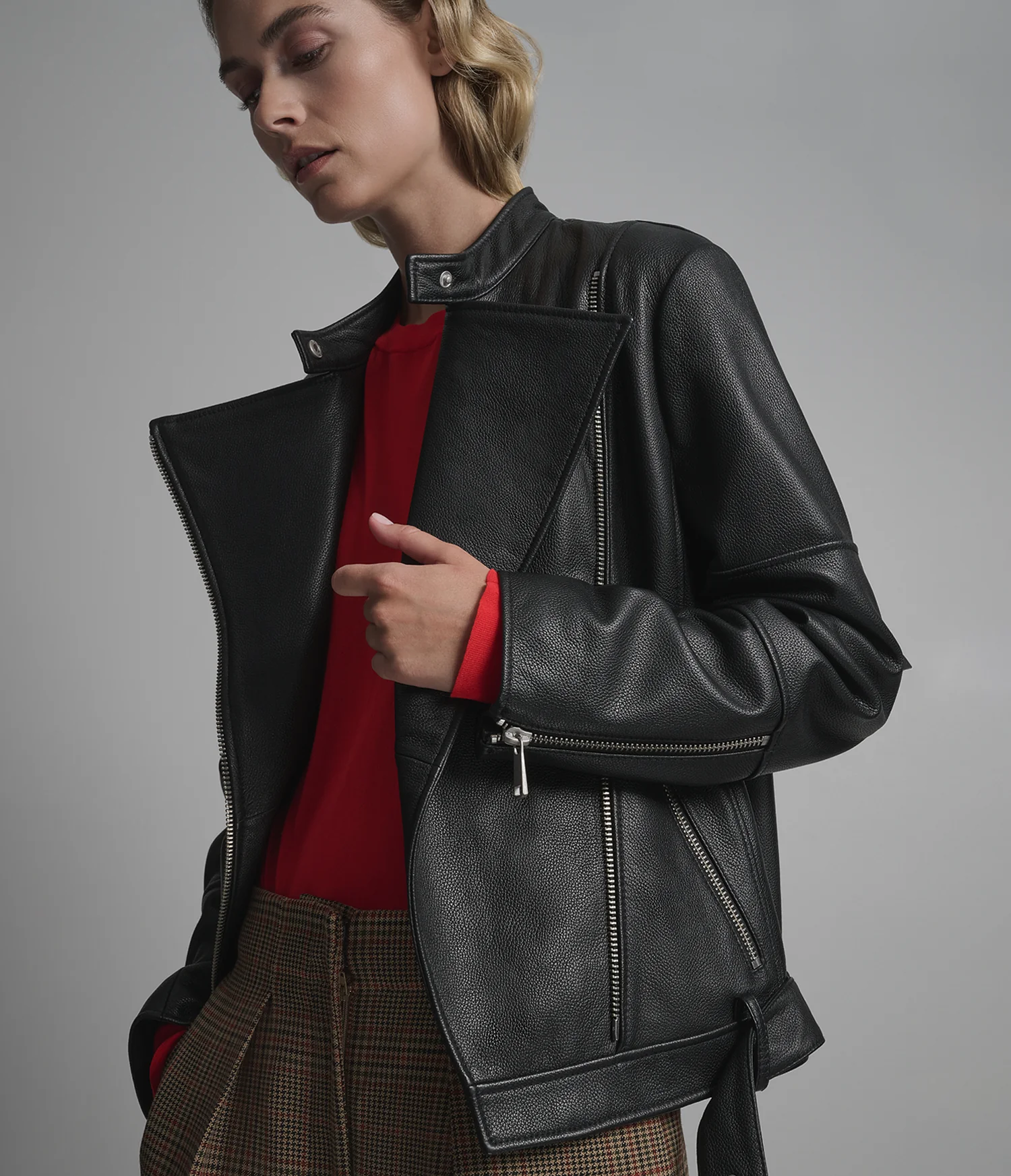 Person wearing a achromatic leather overgarment implicit a reddish apical with plaid pants, posing with overgarment partially open