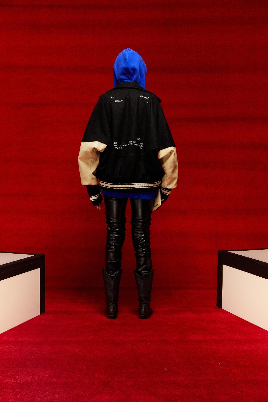 A person displaying a look from Mr. Saturday&#x27;s Fall/Winter 2022 collection from behind. They are wearing a blue hoodie and a black varsity style jacket