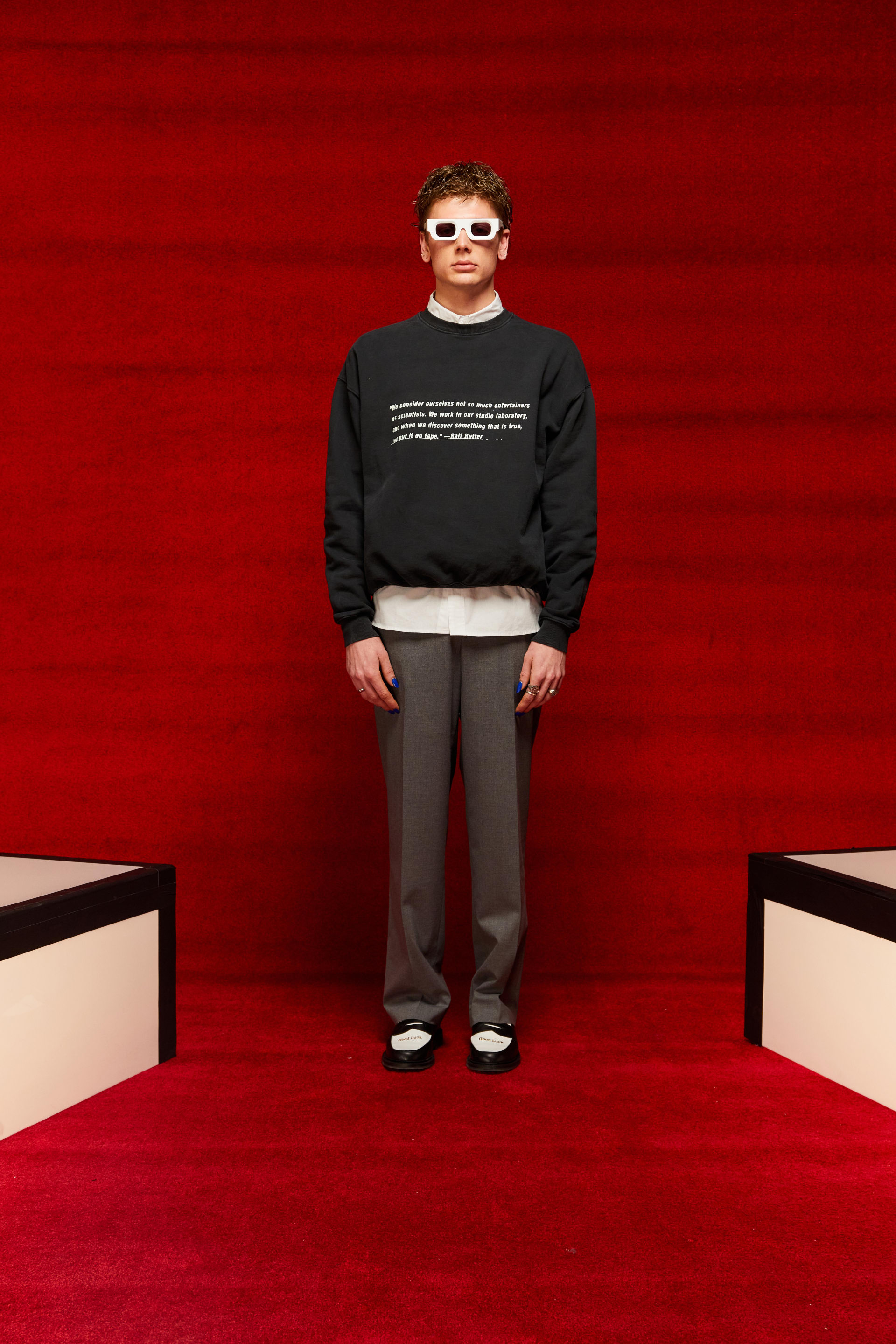 A person displaying a look from Mr. Saturday&#x27;s Fall/Winter 2022 collection. They are wearing white sunglasses, a black crewneck with white text, and grey slacks.