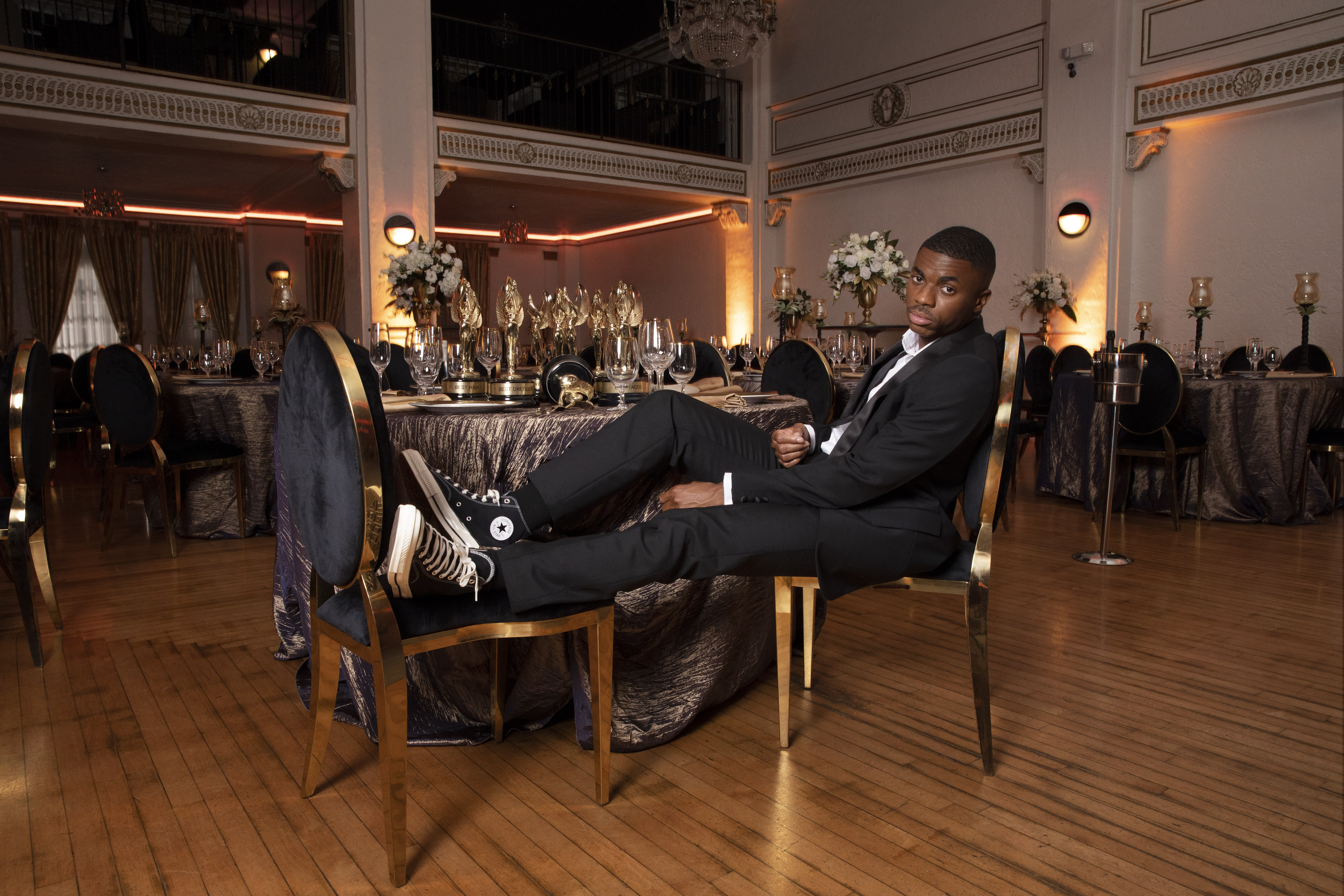 Vince Staples sits on a chair at a table that has numerous trophies on it