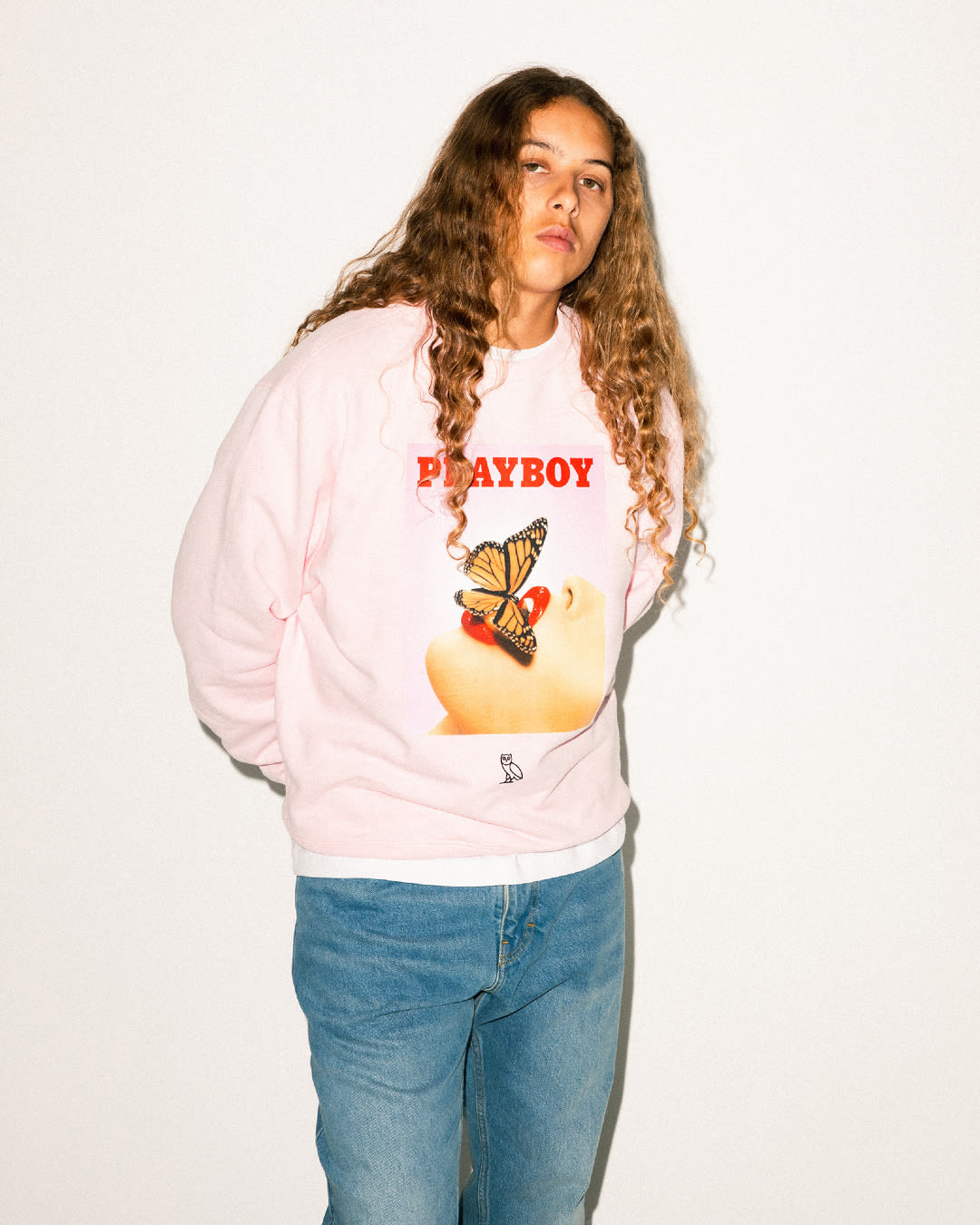 A model wearing a pink crewneck with &quot;PLAYBOY&quot; in red font and an image of a butterfuly on a woman&#x27;s mouth.