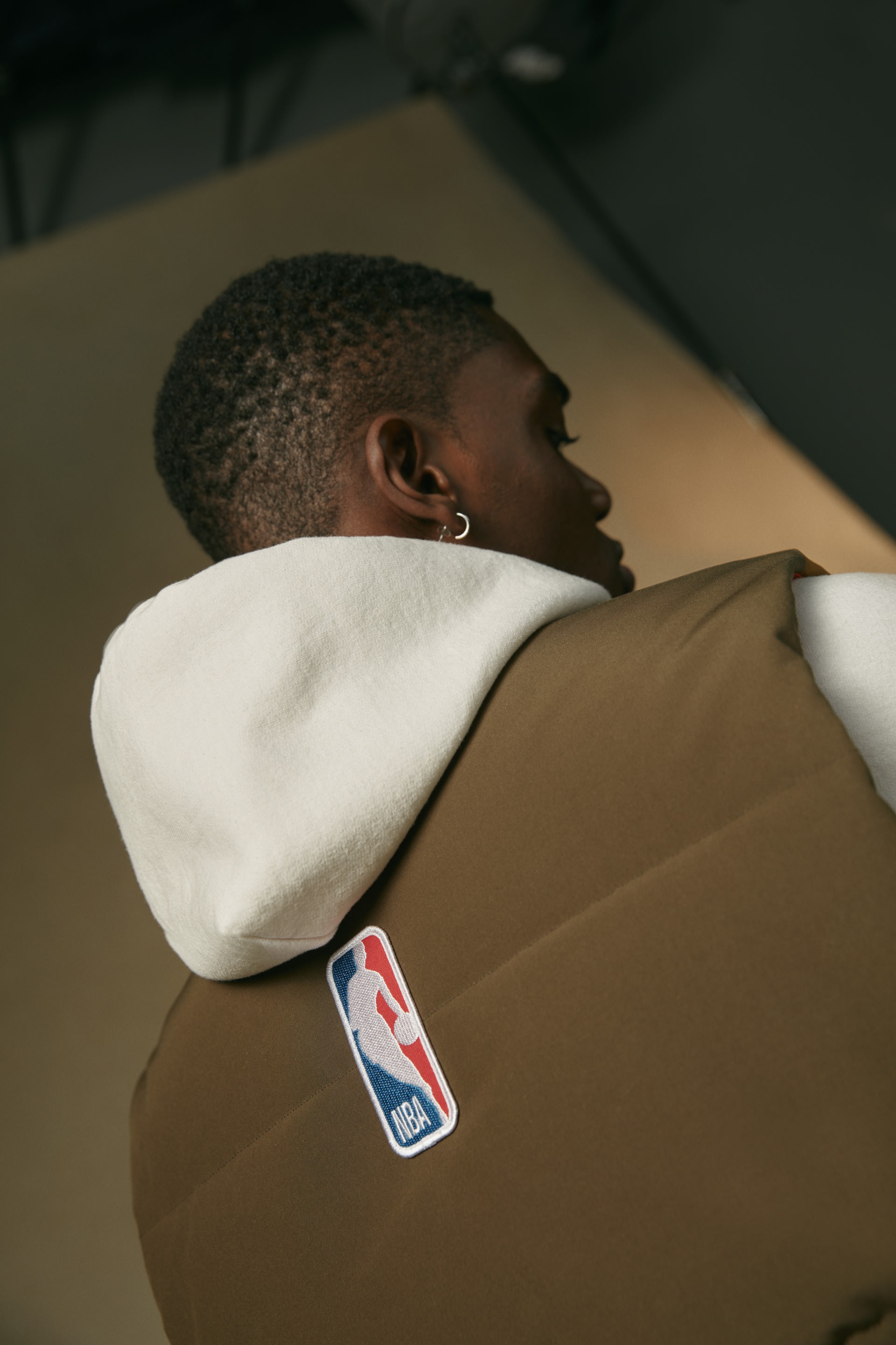 A model wearing a brown Canada Goose vest over a white hoodie from behind, with the NBA logo shown